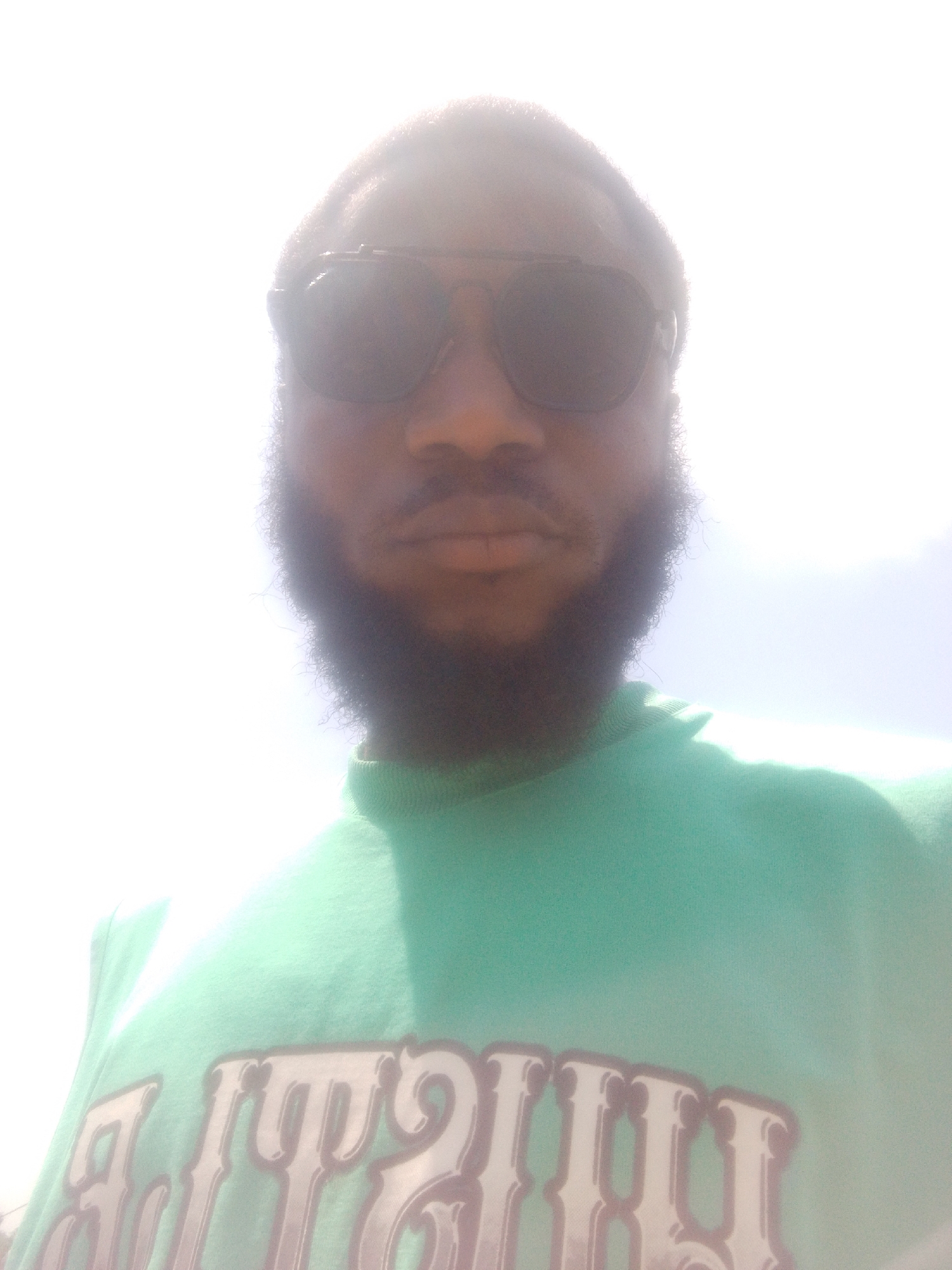 A close-up shot of a dark-skinned man wearing sunglasses and a green t-shirt. The background is overexposed. There is no way to determine the location in Lokoja, Nigeria from the provided picture.