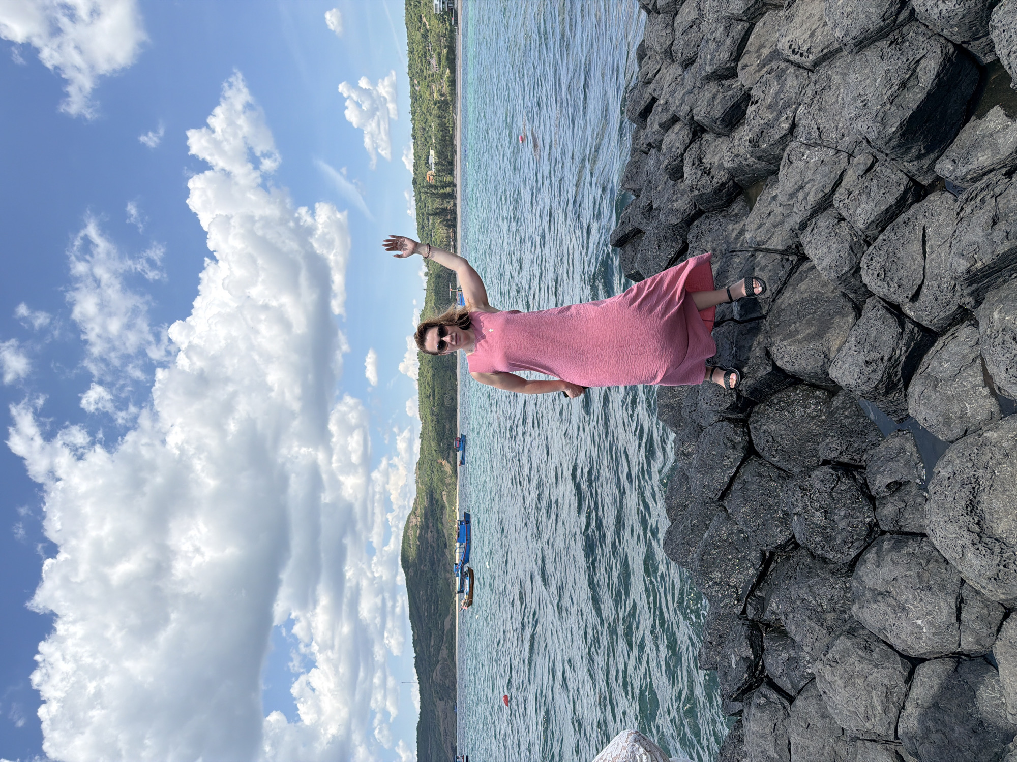 A woman in a long, pink, textured dress and black sandals stands on a rocky shoreline, waving with her right hand. She wears sunglasses. The background features a body of water with several small boats, a green, hilly coastline, and a blue sky with white, fluffy clouds. The rocks are dark gray and irregularly shaped. The scene is consistent with the coastal landscape of Phú Lương, Vietnam.