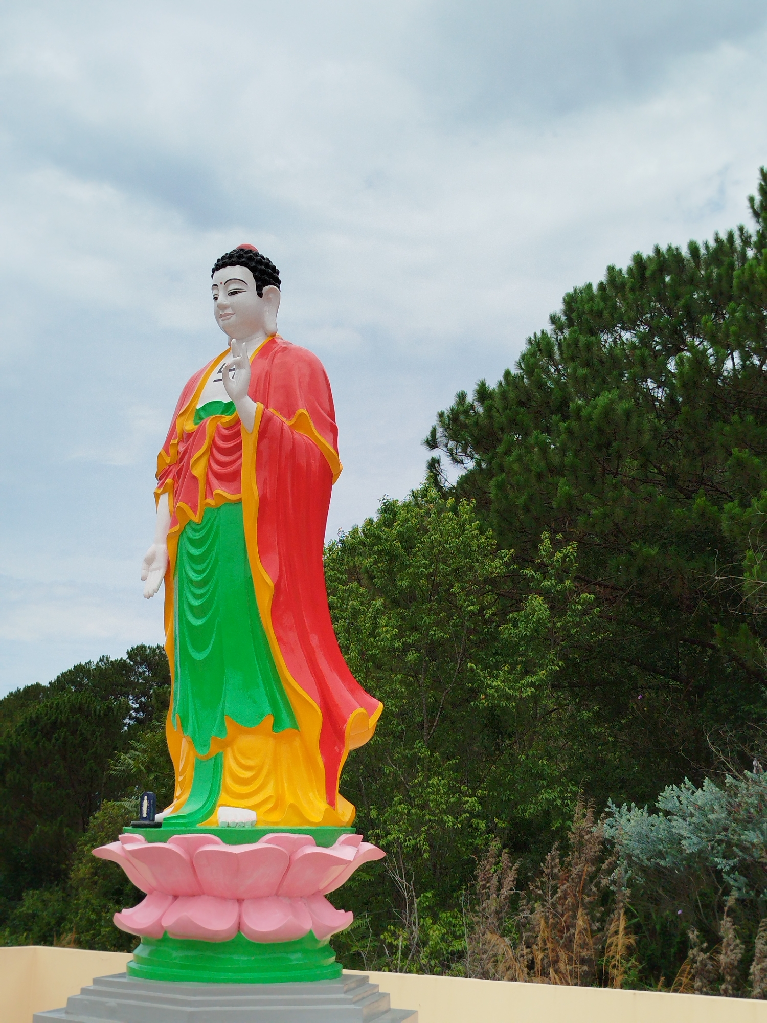 A full, eye-level shot presents a statue of Buddha standing on a large lotus blossom base. The statue is brightly colored, with a white face and hands, a black head of hair, and garments colored red, green, and yellow. The Buddha is posed with his right hand raised in a gesture.
Behind the statue, dense green vegetation forms a backdrop, including a variety of trees and shrubs. The sky above is overcast.
This statue is located in Da Lat, Vietnam.