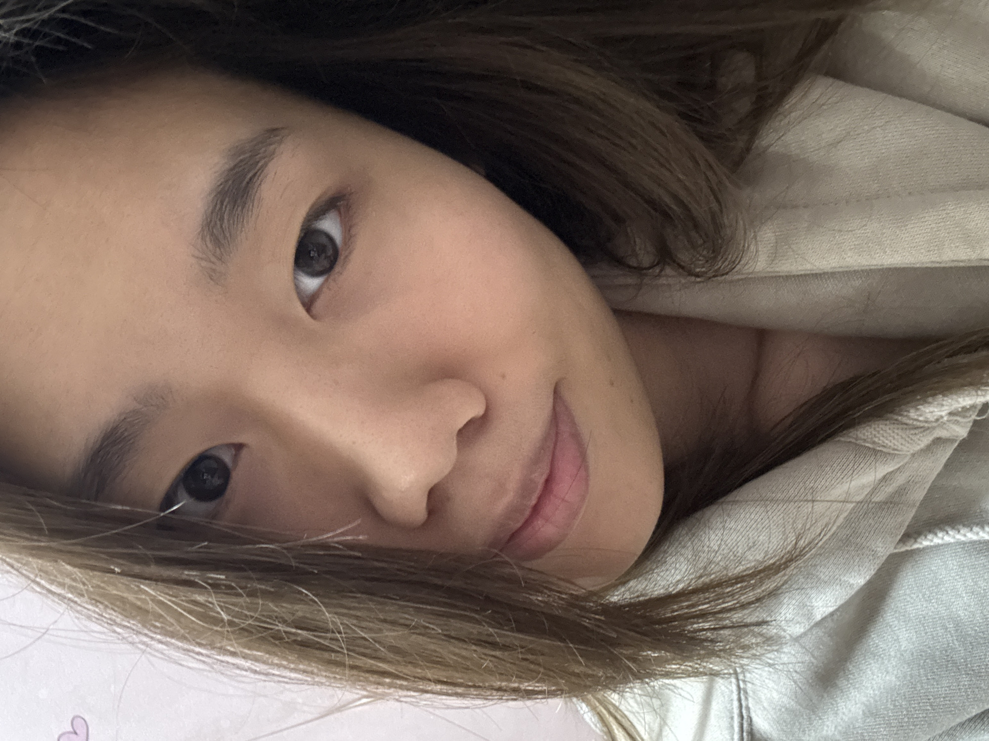 A close-up, angled shot shows a woman's face. She has light brown hair, brown eyes, and fair skin. Part of her hair is in the foreground, covering a portion of the lower left. She is wearing a light-colored hooded sweatshirt. The background is blurry, with what appears to be a pinkish or light-colored surface visible in the lower left corner. There is no readily apparent indication of the location being in the Shapingba District, China.