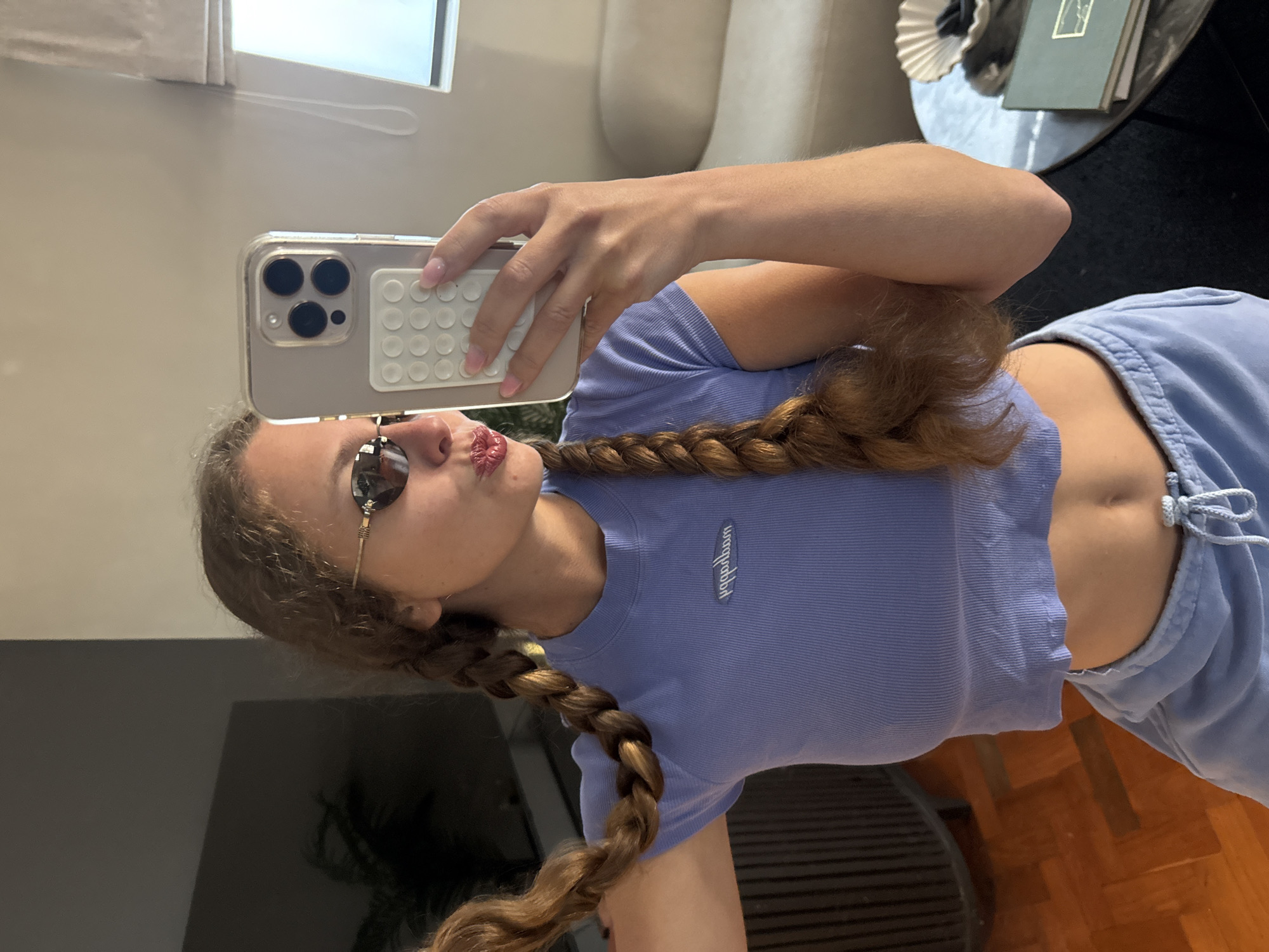 A young woman is taking a selfie in an indoor environment. She's holding a smartphone with a distinctive case featuring circular elements. The woman has long, braided hair, sunglasses, and is wearing a light blue crop top. The background shows a modern interior with a couch, a window with a blind, and a round table with objects on it. There is no information about the location being Cape Town, South Africa in this image.
