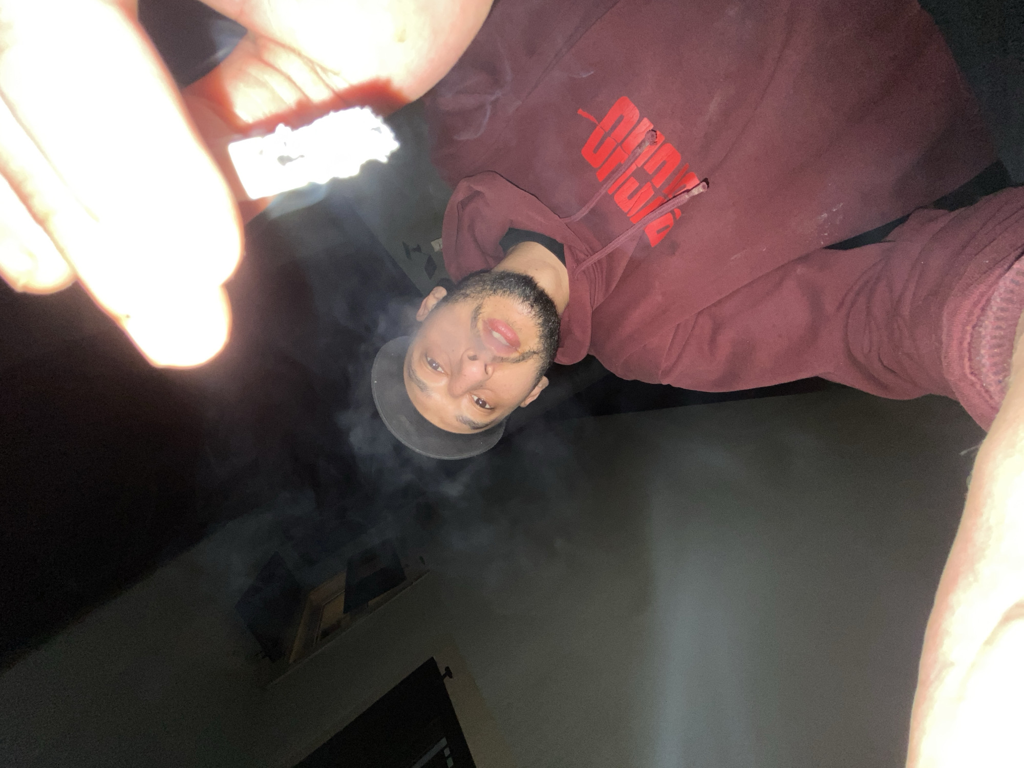 A low-angle shot of a person holding a lit item, possibly a cigarette or joint, in their left hand. The person is wearing a maroon hoodie with red lettering on the chest. The person has a beard and is wearing a dark baseball cap. Smoke is visible near the person's face. The background appears to be an interior space with partially visible walls and a window. The location is not identifiable from the image.