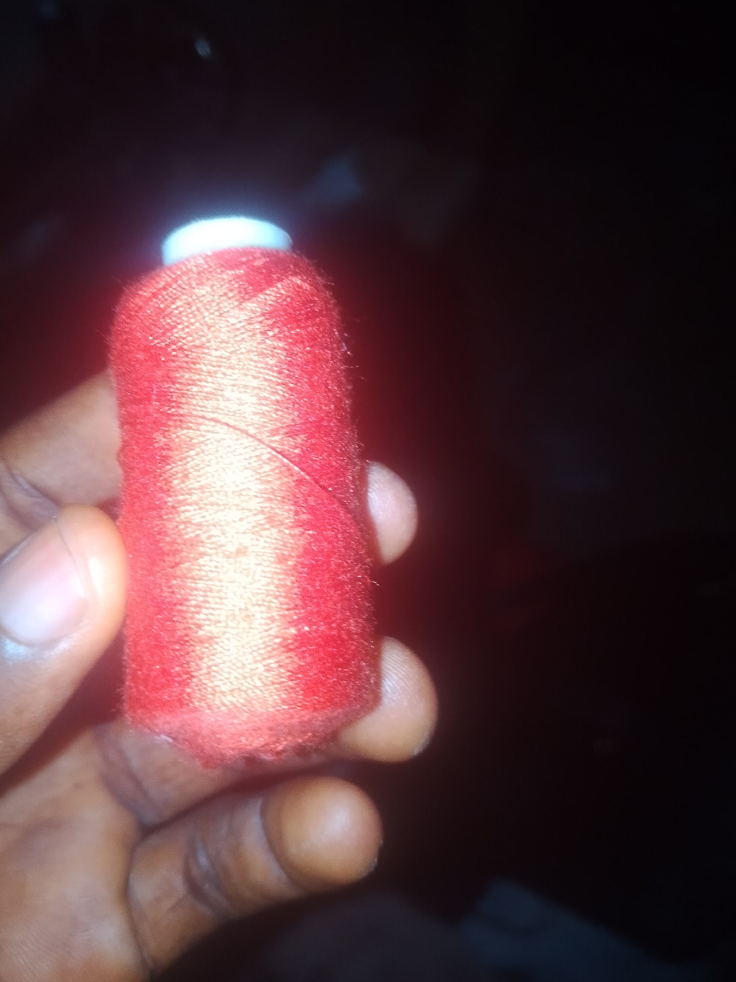 A hand holds a spool of red thread against a dark, out-of-focus background. The spool is cylindrical with a white cap on top. The thread is tightly wound, and a slight sheen suggests it may be made of polyester or silk. The hand holding the spool appears to be that of a person with darker skin tone, with visible fingerprints on the skin. The image is dimly lit, and the background is so dark that it's impossible to discern any specific details about the location. The overall impression is that the photo was taken indoors with limited light, possibly at night. There is no visible text in the image. The setting is likely a domestic environment within Jauro Yinu, Nigeria, given the context. The focus on the thread suggests an interest in sewing or crafting, or perhaps the thread is a necessary item for a task.