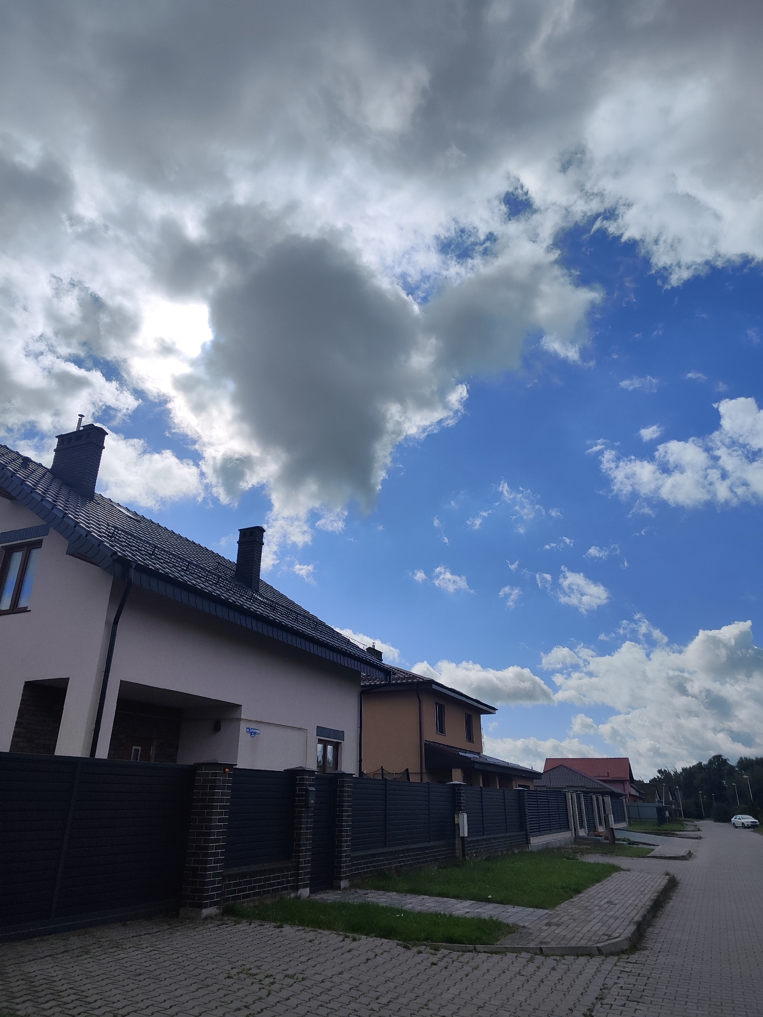 The image shows a street scene in the city of Gvardeysk, Russia, under a cloudy sky with patches of blue. The foreground features a paved walkway and a dark metal fence with brick pillars, separating the street from residential properties. Several houses are visible, with tiled roofs and modern architectural styles. One house in the foreground has a light-colored facade and a dark roof with two chimneys. Further down the street, other houses with different colored roofs and facades are visible, along with a tree line in the background. A white car is parked on the right side of the street, which is also paved. The scene appears to be a quiet suburban or residential area.