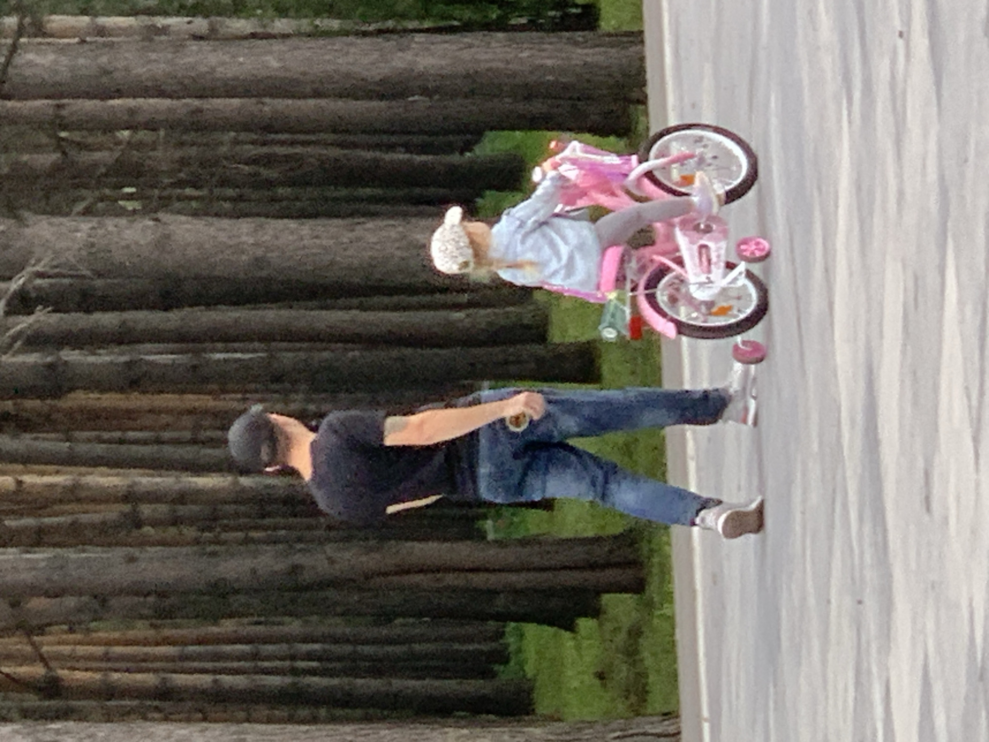 A man wearing a black t-shirt, blue jeans, and a dark baseball cap walks on a paved path in a park. He is holding a small object in his right hand. To his right, a young girl with blonde hair, wearing a light blue jacket, grey pants, and a white patterned cap, rides a pink bicycle with training wheels. The bicycle has a basket on the front and a small green object attached to the back. They are in a wooded area with tall, dark trees in the background. The ground is a light-colored paved surface. The image appears to be taken in the city of Prokopyevsk, Russia.