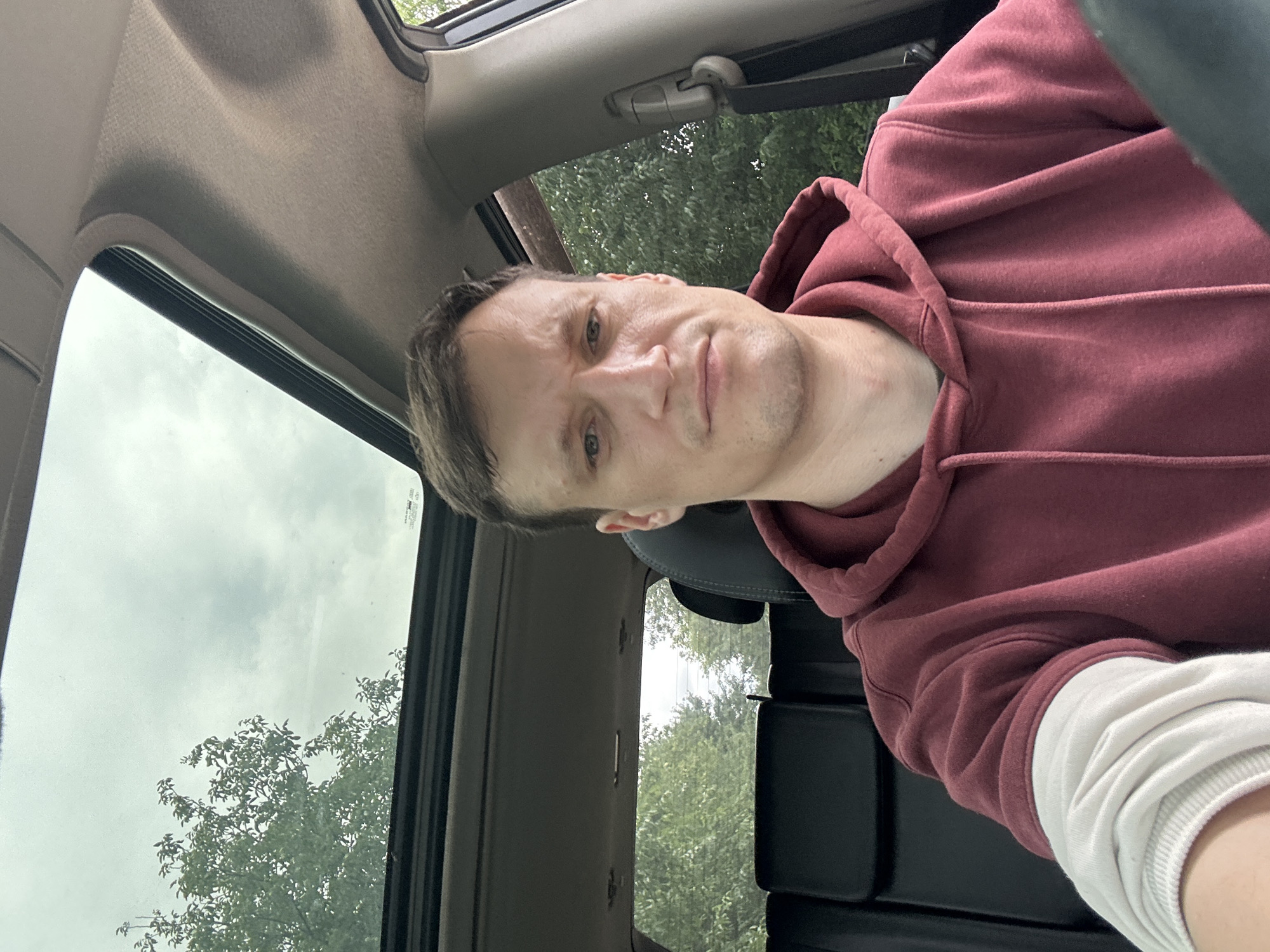 A man is pictured from the chest up, inside a vehicle. He is looking directly at the camera with a neutral expression. He is wearing a dark red hooded sweatshirt over a white long-sleeved shirt. The interior of the vehicle is visible, including a dark-colored seat and the headliner. The vehicle's sunroof is open, revealing a cloudy sky and trees outside. The scene appears to be outdoors, possibly on a street in Korchivka, Ukraine, given the greenery and the style of the vehicle.