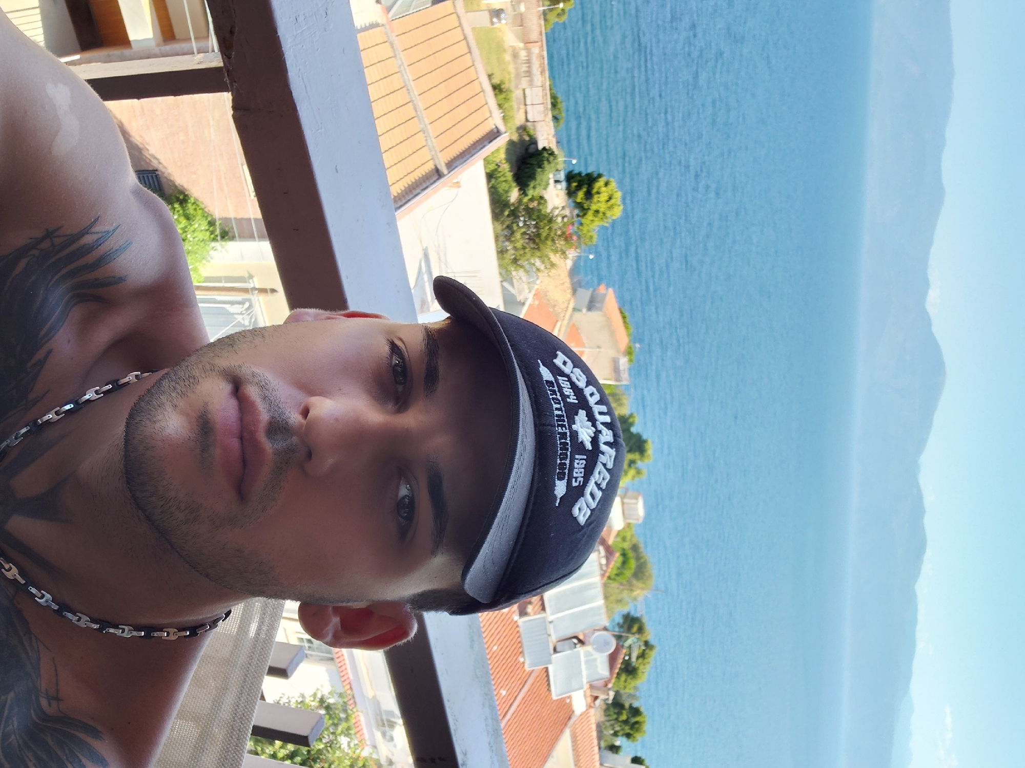 A man wearing a black baseball cap with white lettering, is pictured in the foreground. He has dark hair and is looking directly at the camera. He is shirtless and has tattoos on his chest and shoulder. He is wearing a silver chain necklace. Behind him, there is a balcony railing. In the background, there are buildings with orange tiled roofs, green trees, and a body of blue water. Further in the distance, there are hazy mountains under a clear blue sky. The location is the Municipal Unit of Aegio, Greece.
