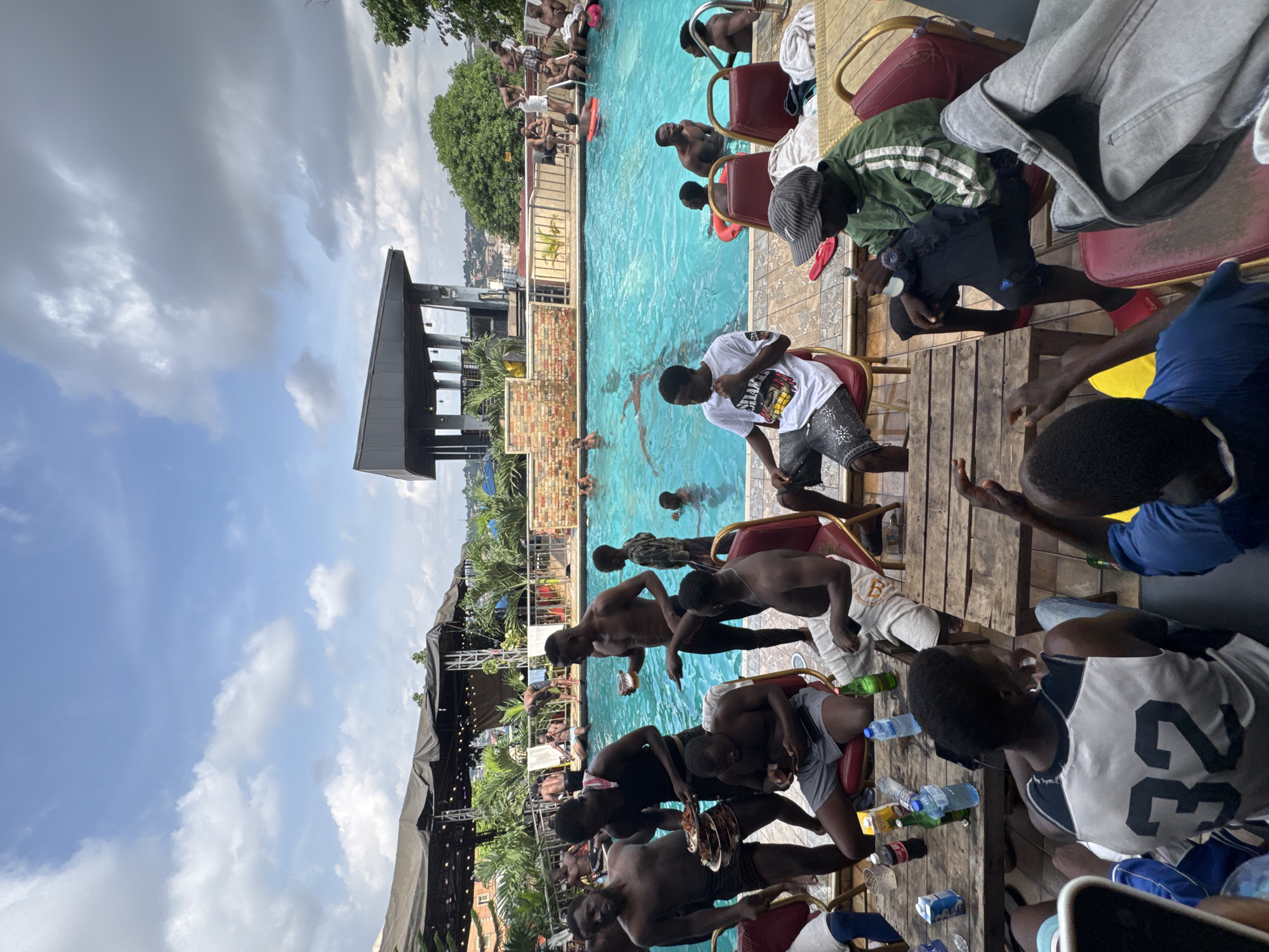The image depicts a lively gathering around a swimming pool, likely at a resort or club in Nkpor, Nigeria. The scene is bustling with activity, as a group of men, mostly young adults, enjoy leisure time. Several individuals are in the bright blue swimming pool, some swimming, others standing and conversing. Others are seated at wooden tables and chairs positioned around the pool deck, engaged in conversation and consuming food and drinks.
Objects visible include plates of food, bottles of beverages (including what appears to be Coca-Cola and water), a smartphone, and towels. The architecture suggests a rooftop setting, with a building structure visible in the background and a large tented area with lights. Palm trees and other greenery are interspersed around the pool area, adding to the tropical ambiance. The sky is partly cloudy, indicating daytime. The overall atmosphere is one of social enjoyment and relaxation.