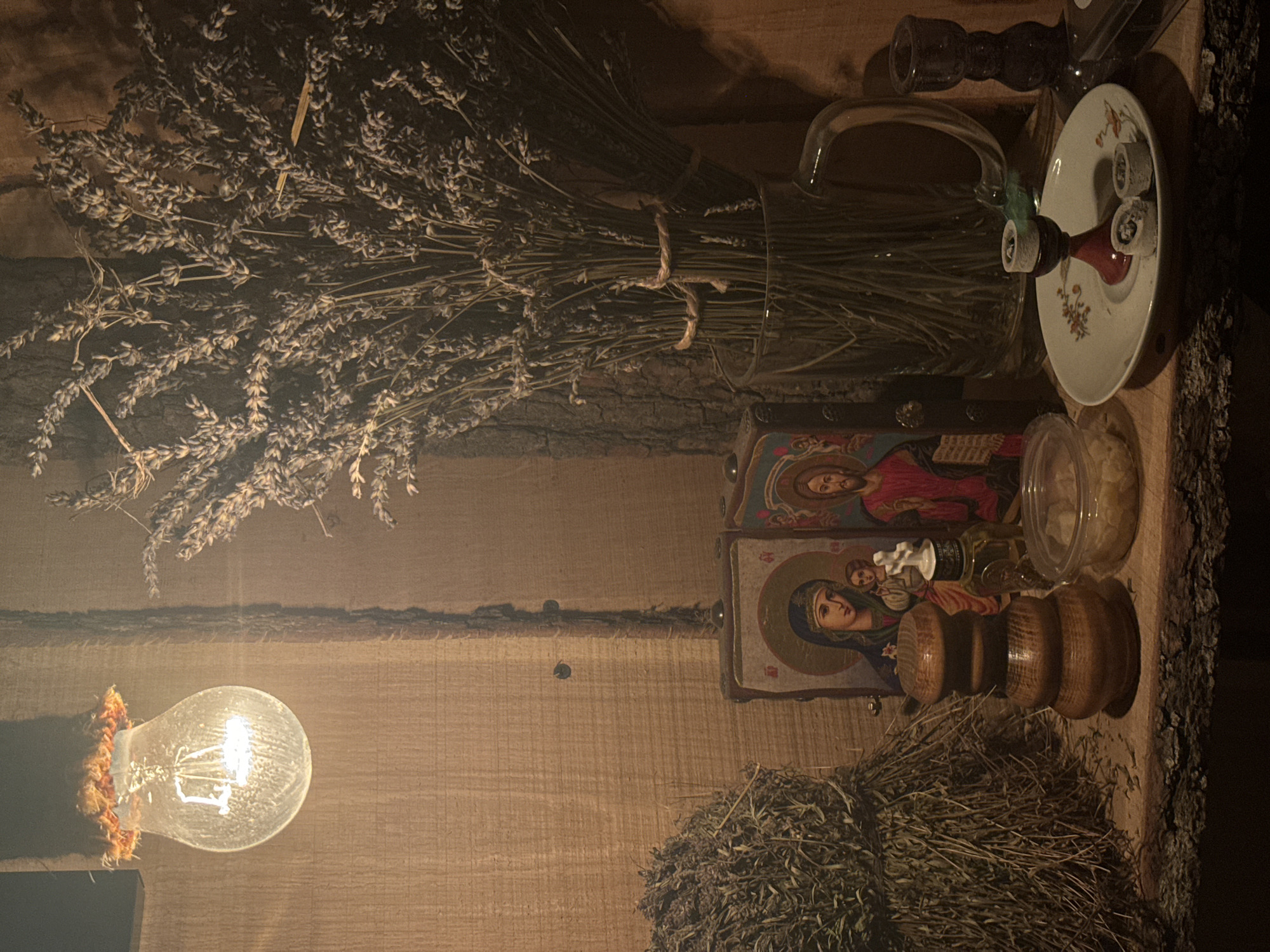 The image depicts a dimly lit, rustic interior, likely a domestic setting. A lit incandescent light bulb is visible in the lower left corner. Above it, a rough-hewn wooden surface serves as a shelf. On the shelf, there are several items: a large bunch of dried lavender tied with string, a glass pitcher, a small plate holding burning incense cones, two religious icons depicting Orthodox figures, a small transparent container, and a wooden object resembling a mortar and pestle. The overall atmosphere is one of tranquility and spirituality. Based on the inclusion of religious icons it is highly likely this location is in an Eastern European city, based on the prompt provided, this location is in Belgrade, Serbia. There are no people visible.