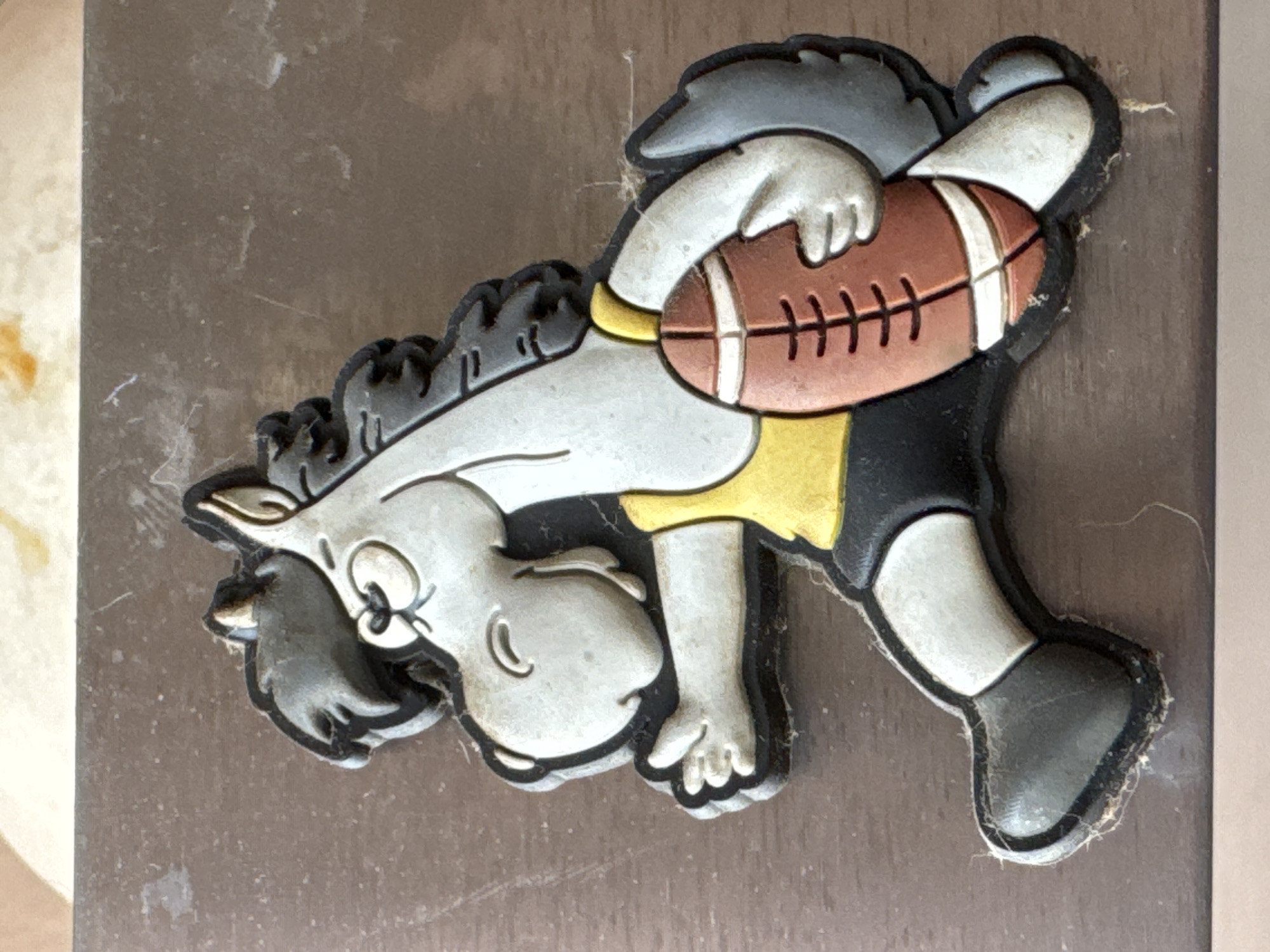 A cartoon horse mascot, likely associated with a sports team, is depicted in a dynamic pose, holding a football. The mascot is gray with black mane and tail, wearing a yellow tank top and black shorts. It has a determined expression with closed eyes and a slightly open mouth. The football is brown with white laces and stripes. The mascot is placed on a textured, brown surface, possibly a table or countertop, in an indoor setting. The background is out of focus, but appears to be a light-colored, possibly food-related item. The image does not provide any information about its location in Bishkek City, Kyrgyzstan.