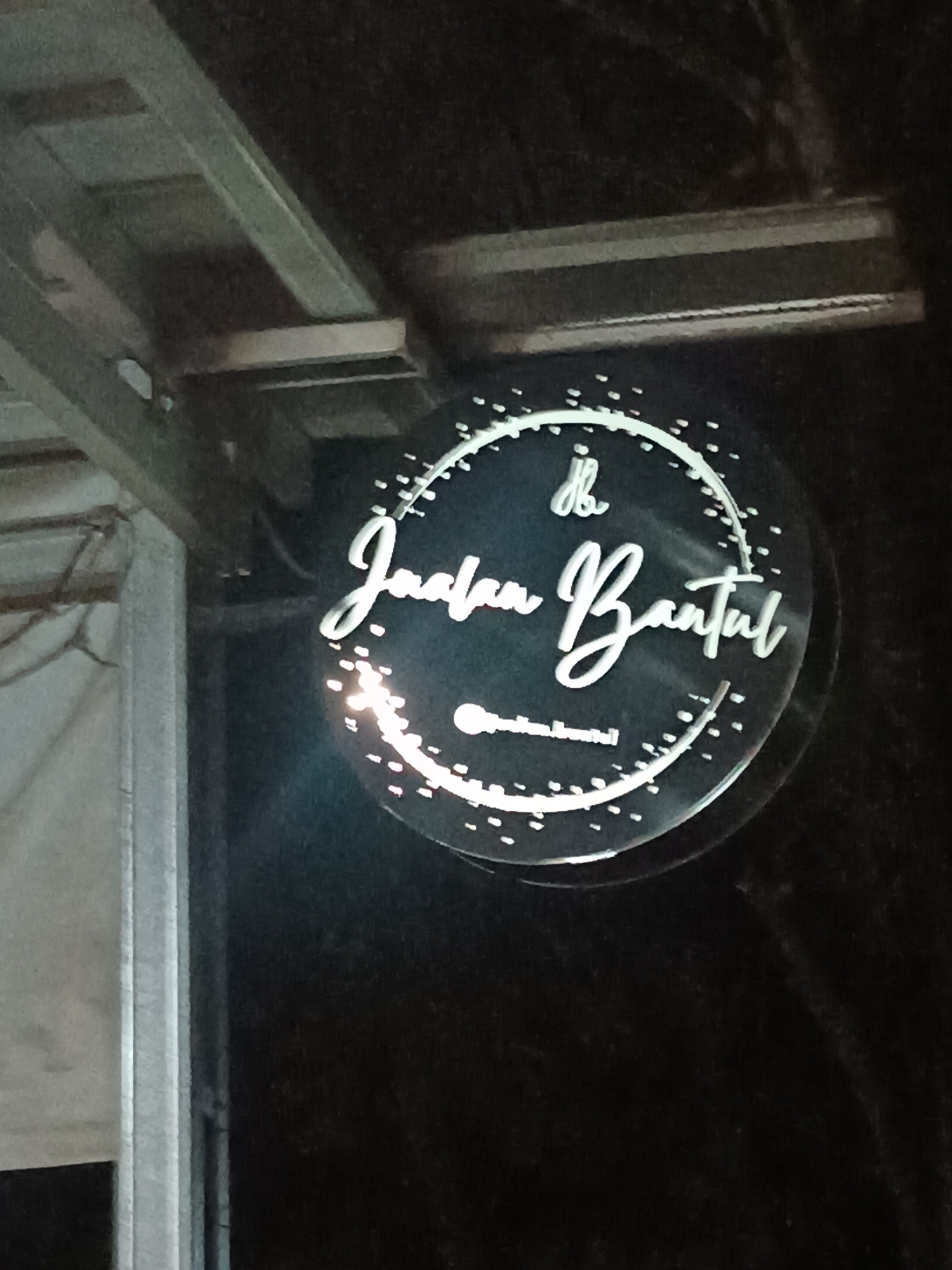 A circular, illuminated sign reads "Jalan Bantul" in white cursive lettering. The sign is suspended from a metal structure, likely part of an overhang or building facade. Small, scattered white lights adorn the perimeter of the circle, creating a decorative effect. Below the main text, a smaller line of text, possibly a social media handle, is visible. The sign is set against a dark background, suggesting it is nighttime. The overall scene appears to be an outdoor location in Jetis, Indonesia, likely a street-level business or establishment.