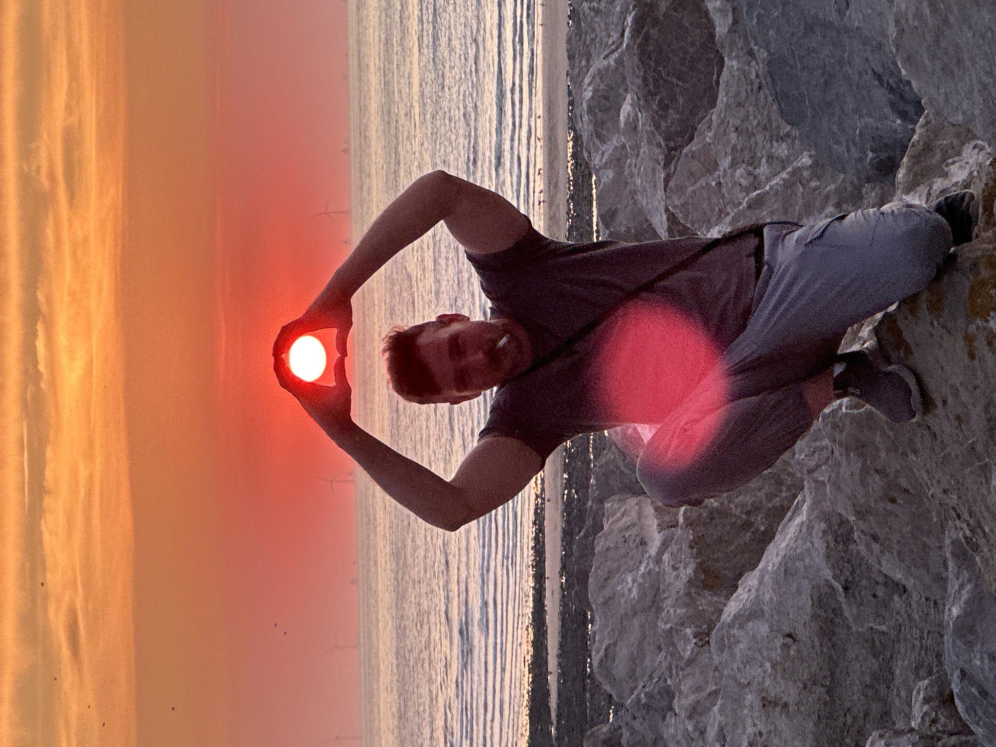 The image depicts a man squatting on a rocky shoreline with the sea and a sunset in the background. The man is holding his hands up to frame the setting sun, which appears as a bright red orb. He is wearing a dark t-shirt, grey cargo pants, and dark athletic shoes. A cigarette is visible in his mouth. In the distance, several wind turbines can be seen on the horizon, suggesting a coastal location. The sky is filled with warm hues of orange and red, characteristic of a sunset. The foreground consists of large grey rocks. This scene is identifiable as the shoreline of Le Tréport, France, known for its distinctive rock formations and the presence of offshore wind farms.