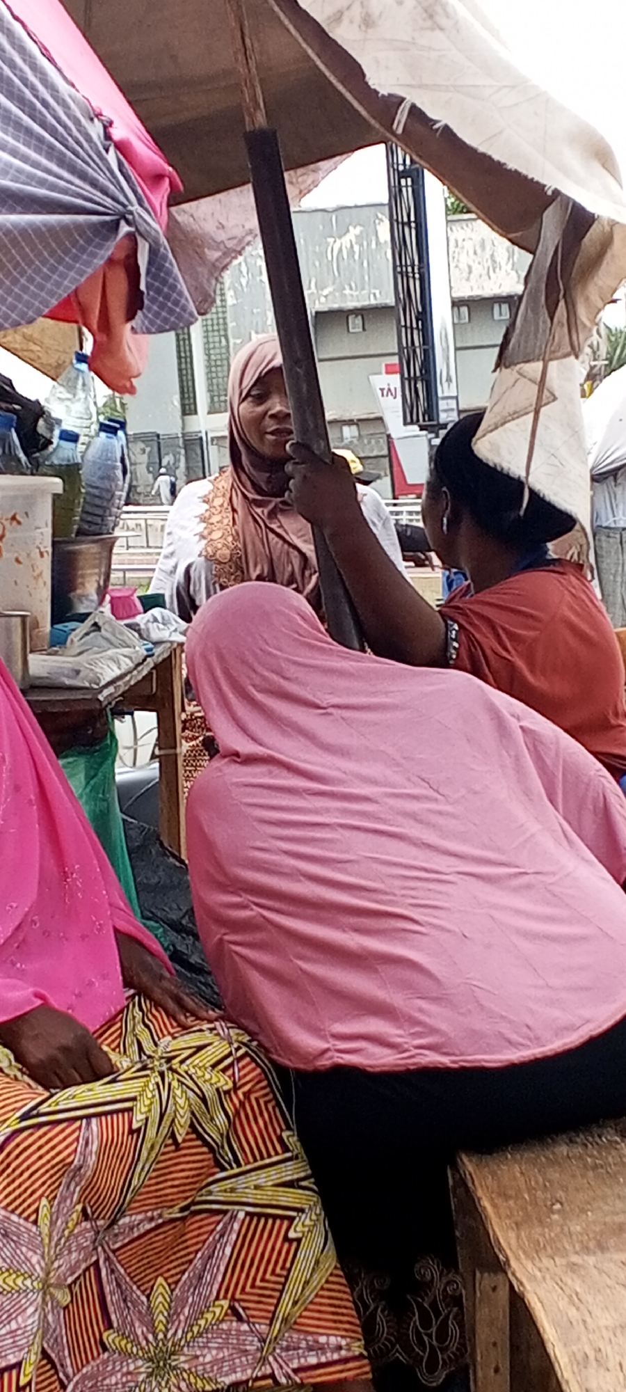 Three women are gathered beneath a makeshift canopy in Maiburiji, Nigeria. One woman is facing forward, wearing a brown hijab and a white shirt with embroidered detailing. Another woman, dressed in a red shirt, has her back to the camera, her hand resting on the canopy's pole. The third woman, wearing a pink hijab, is seated on a wooden bench next to a woman in a pink shirt and a brightly patterned skirt. Water bottles and other items are visible on a table nearby. A building with the letters "TAJ" is in the background.