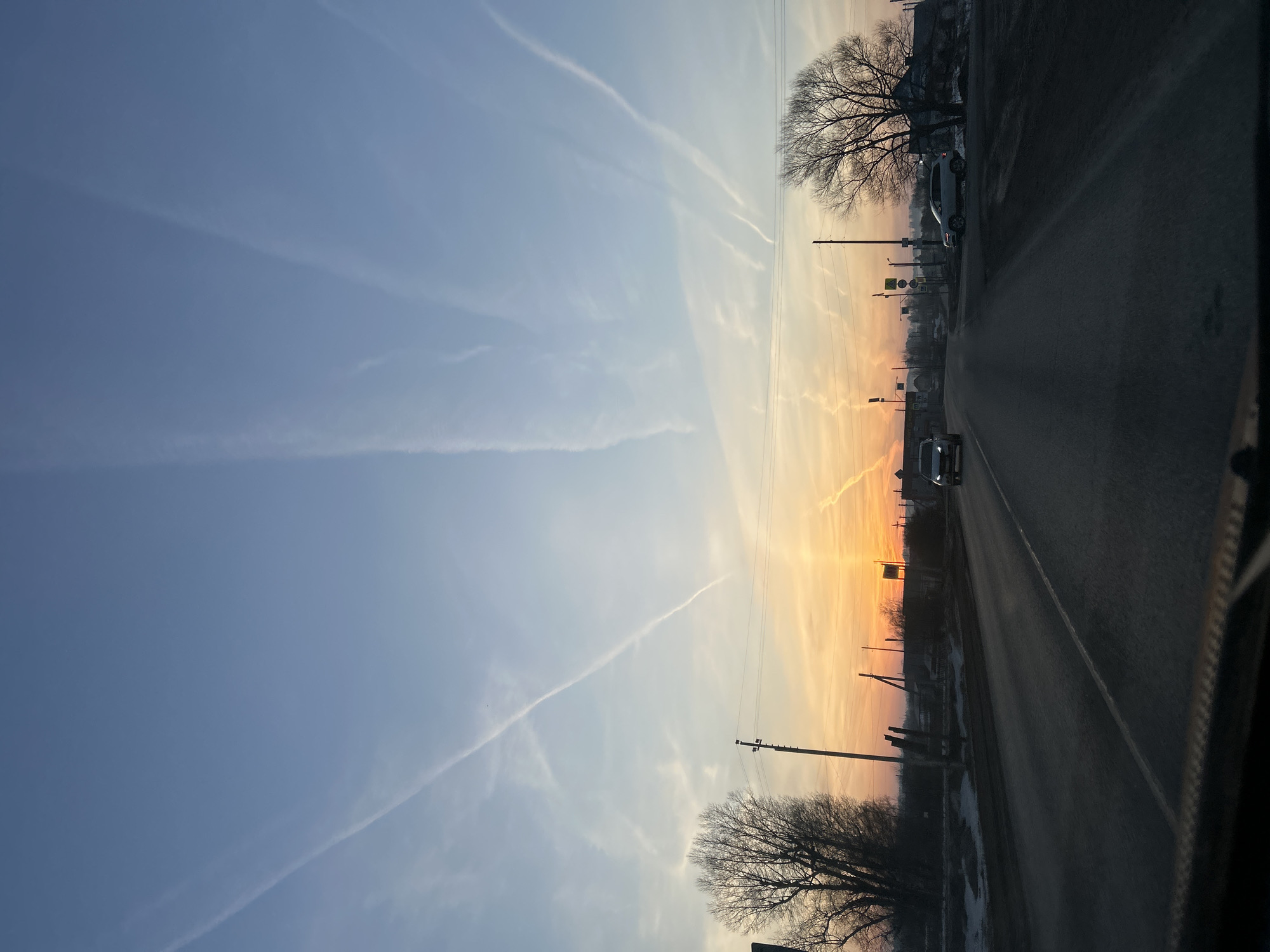 The image depicts a road leading into the sunset in what appears to be a rural area. The view is likely taken from inside a moving vehicle, as the bottom right edge shows part of the car's interior. On either side of the road are bare trees, typical of winter or early spring. Power lines run alongside the road. The sky is predominantly blue with contrails of aircraft. The setting sun creates a gradient from yellow to orange near the horizon. There is a vehicle on the road ahead, and another parked on the side of the road, near a bus stop. There is also some remaining snow beside the road. Based on the location Гаревая, Russia, the weather is likely cold.