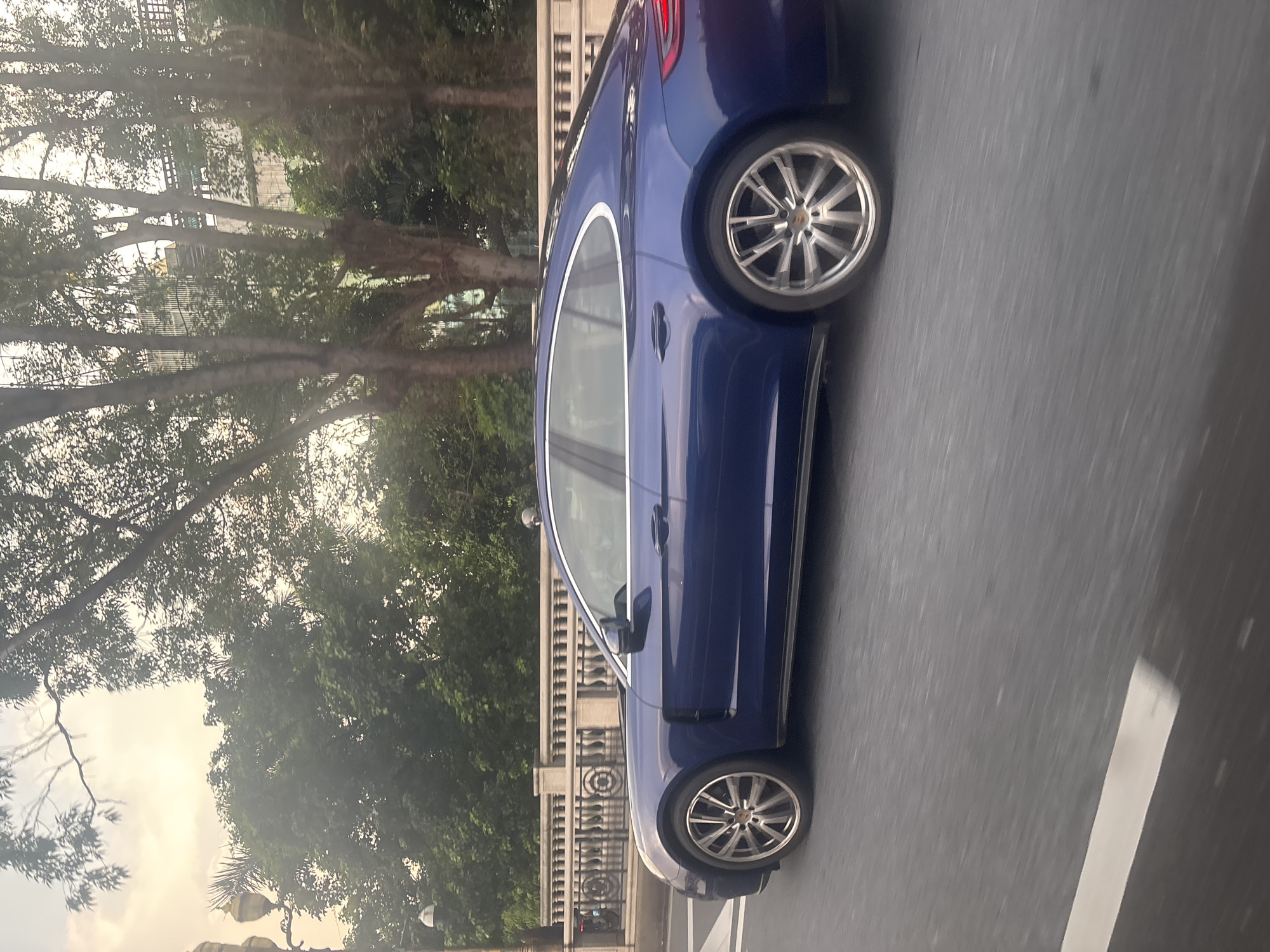 A blue Porsche Panamera is captured in a motion blur, traveling on a road in Haizhu District, China. The car is seen from its side profile, showcasing its sleek design and alloy wheels. In the background, a stone bridge with decorative railings spans across, and lush green trees with a clear sky overhead create a serene backdrop. The image conveys a sense of movement and urban landscape.