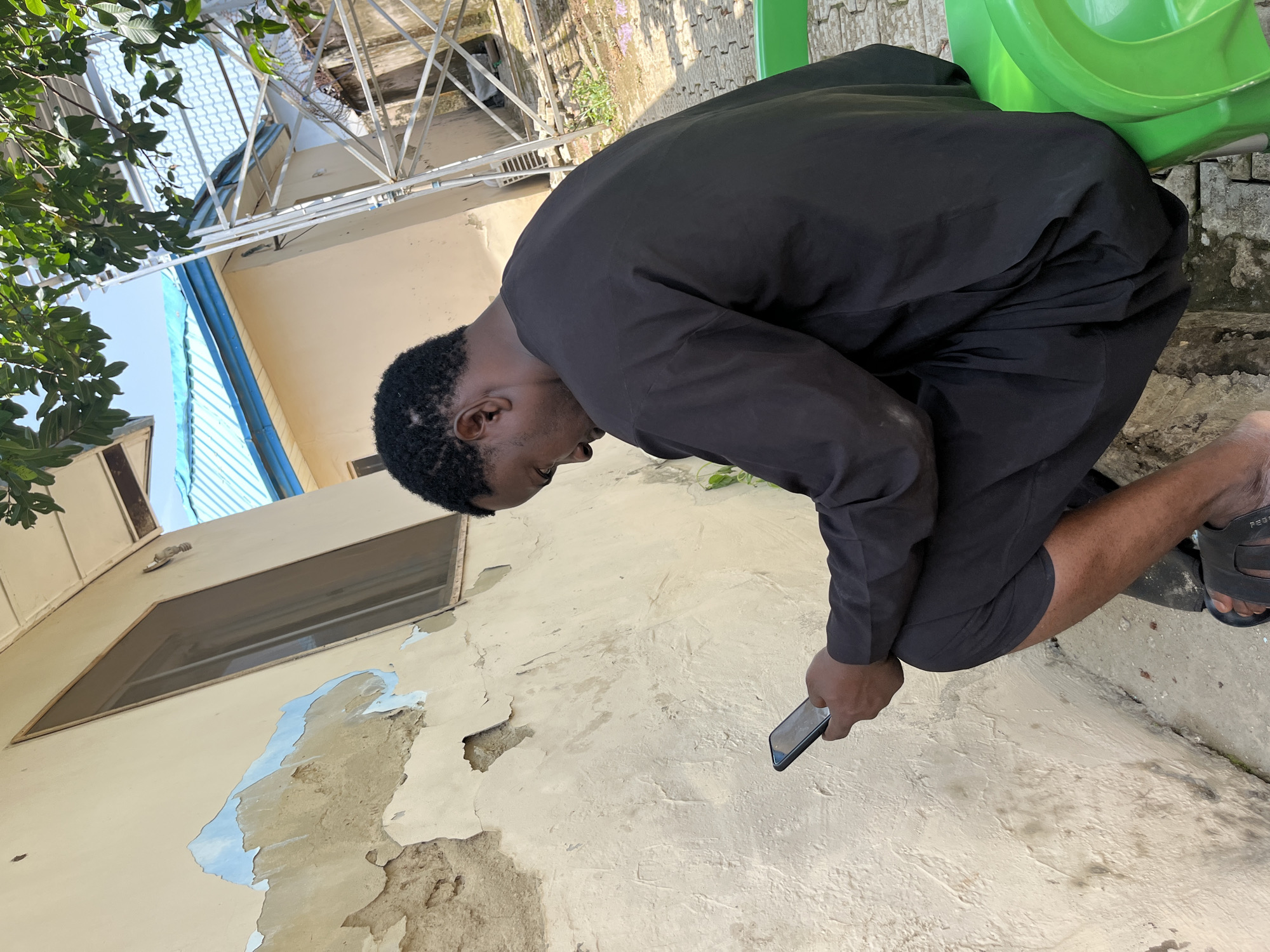 The image depicts a young man in Galadimawa Village, Nigeria. He is wearing a black traditional outfit and dark shorts, with black sandals on his feet. He is leaning over, looking down at his mobile phone, which he holds in his right hand. The setting appears to be an outdoor area with a weathered, light-colored wall behind him. The wall shows signs of peeling paint, revealing patches of blue underneath. To the left of the man, there is a window with a wooden frame. Above and behind the wall, a metal structure and some greenery are visible, suggesting a semi-urban or rural environment. A bright green object, possibly a plastic chair, is partially visible in the upper right corner of the frame. The lighting suggests it is daytime, and the weather appears to be clear. The man's posture and focus on his phone indicate he is engaged in using the device. There is no visible text in the image.