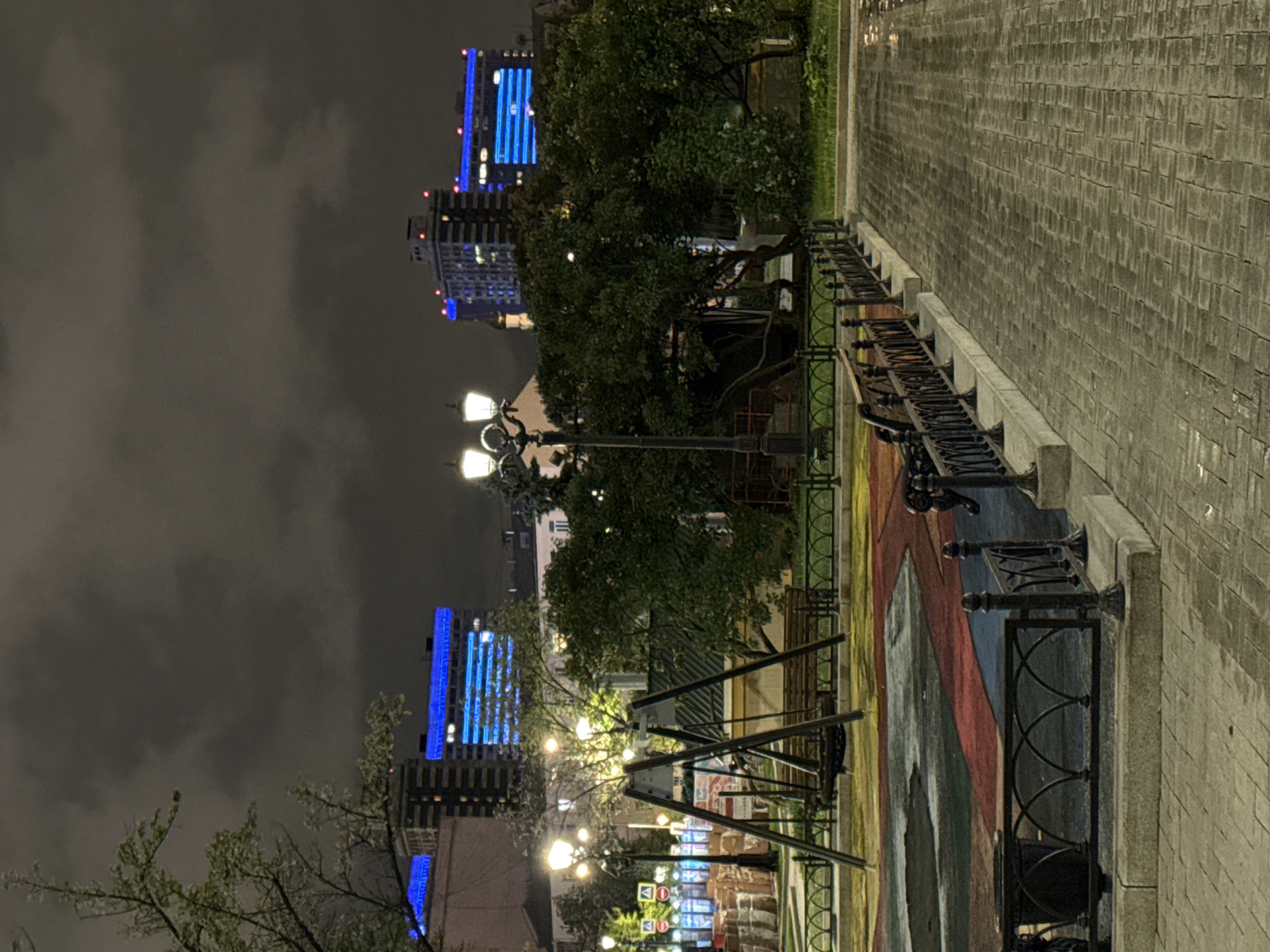 The image shows a night scene in Moscow, Russia. In the background are tall buildings with blue neon lights illuminating their facades. A line of illuminated streetlights leads into the distance. There is a paved sidewalk on the right, and a park-like area with trees, benches, and a metal fence on the left. The sky is cloudy and dark. There are no people visible. The overall scene is urban and atmospheric.