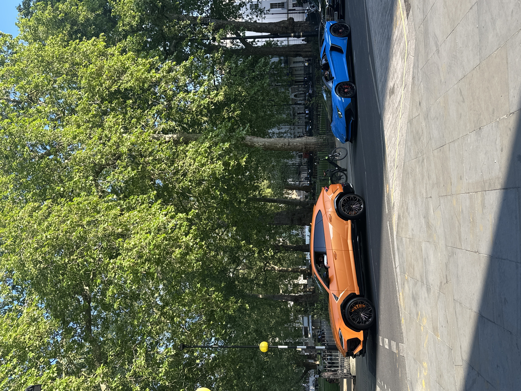 This image captures a vibrant street scene in London, United Kingdom, under bright daylight. Two luxury sports vehicles dominate the right side of the frame: an orange Lamborghini Urus SUV in the foreground and a blue Lamborghini sports car positioned behind it. Both cars feature distinct black multi-spoke wheels. The blue car displays a license plate reading "P23 UAN".
The left side of the image is filled with lush, green foliage of mature trees, likely London plane trees, indicative of summer. These trees line the street, partially obscuring the traditional light-colored buildings in the background. The street itself is paved, with a visible double yellow line marking the curb. Several bicycles are parked on the side of the road, near the blue car. Strong shadows are cast across the pavement, suggesting a sunny, clear day, likely midday or early afternoon. A black street pole with a prominent yellow spherical marker is also visible near the orange car. The perspective is slightly elevated, offering a view down onto the street and its luxurious occupants.