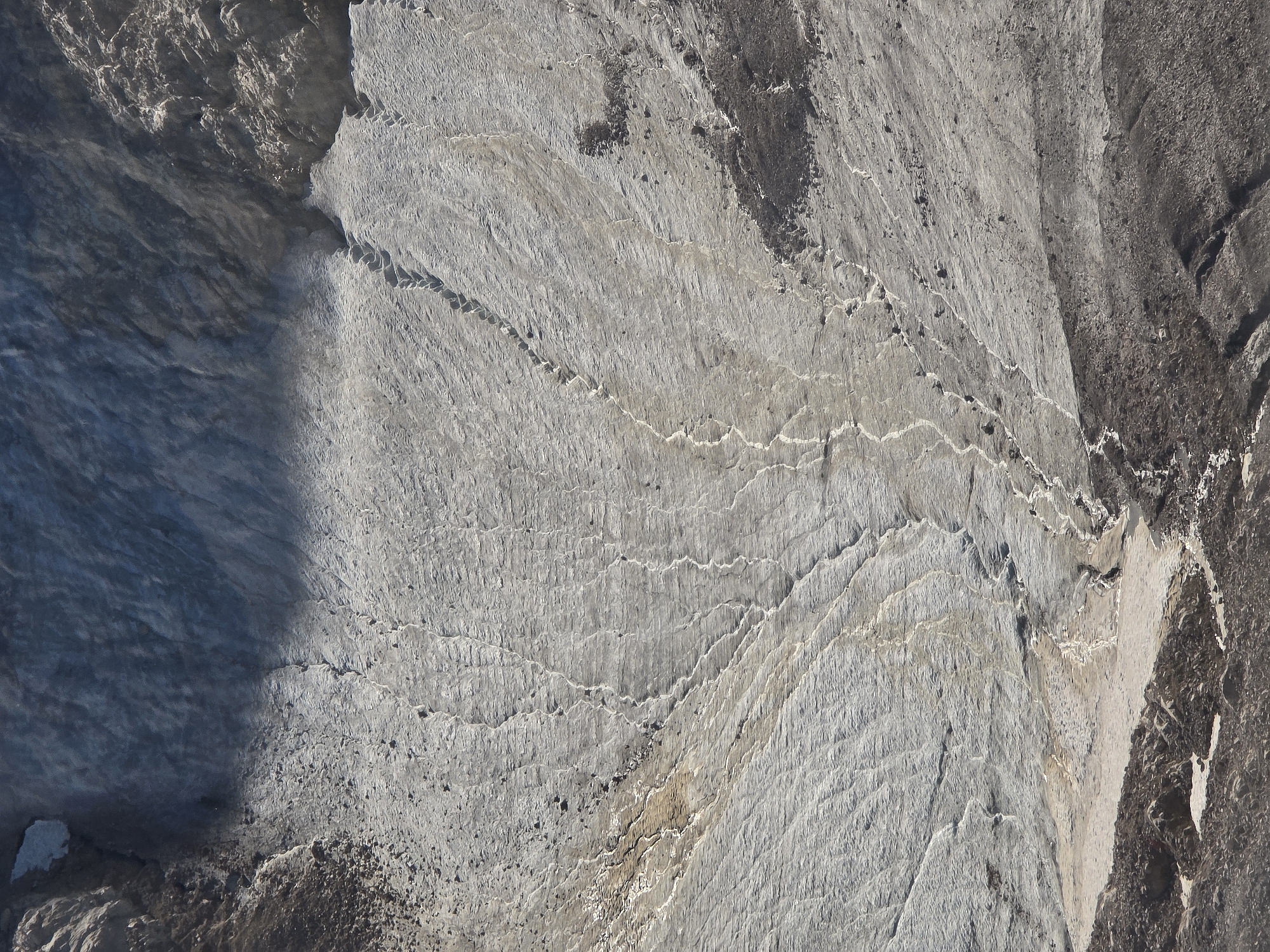 The image depicts a close-up, high-angle view of a glacier, likely part of the Zugspitze massif near Garmisch-Partenkirchen, Germany. The glacier surface is predominantly a light grey, textured with numerous parallel striations and finer, branching channels indicative of meltwater flow. Scattered dark rocks and debris are embedded within the ice. The upper portion of the glacier exhibits a darker, bluer hue, suggesting denser, older ice. The texture of the ice varies, with smoother areas and rougher, more fractured sections. The overall impression is one of a vast, ancient, and dynamic icy landscape.