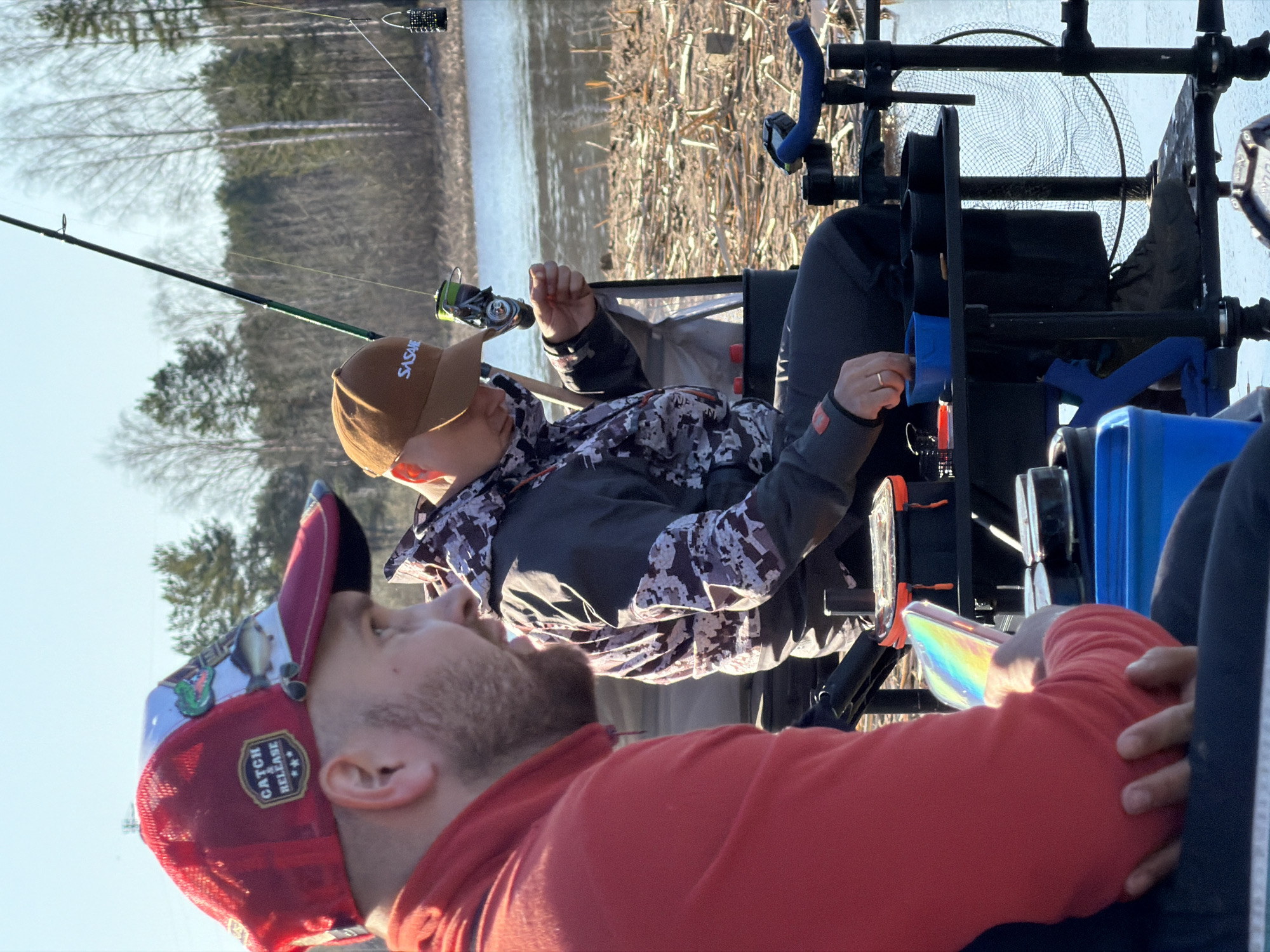 Two men are fishing on a body of water. One man in the foreground wears a red sweatshirt and a multi-colored baseball cap with a "Catch Release" emblem. The man in the background wears a brown cap with the word "SASAME" on it, a camo jacket, and is actively fishing with a rod and reel. A black fishing setup with blue components is visible between them. The backdrop shows water, reeds, and bare trees. The location is likely Chaikovsky, Russia, based on the cap’s brand.