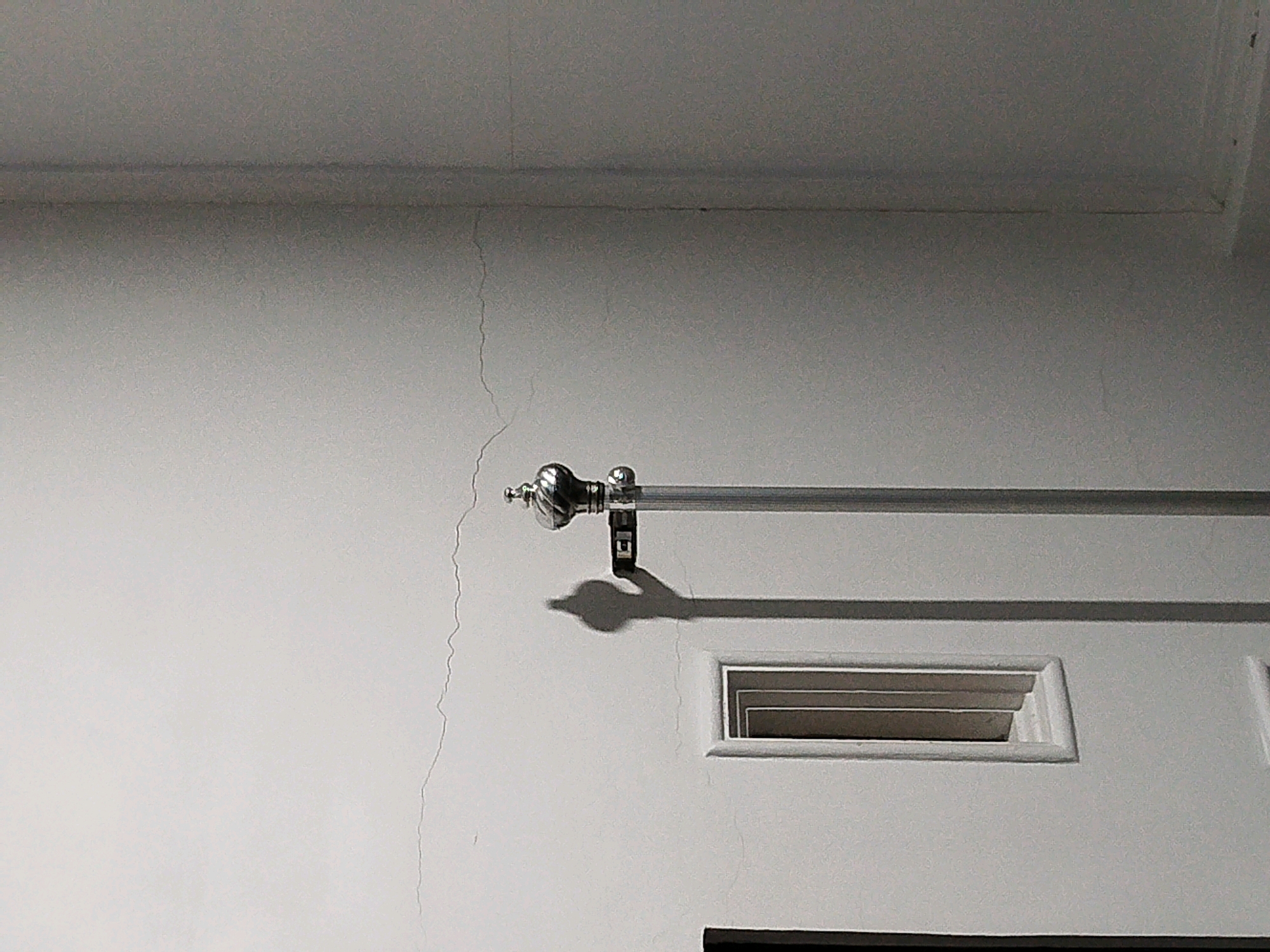 The image shows a close-up view of a light-colored wall with a horizontal metal curtain rod mounted on it. A curtain rod bracket is visible, casting a shadow on the wall. Below the curtain rod, there is a rectangular vent or window opening with a white frame. Several thin, vertical cracks are visible on the wall's surface, extending from near the top of the frame down towards the bottom of the image. The scene appears to be an interior shot, likely in a residential or commercial building, with a neutral, textured wall. The lighting suggests an indoor environment with artificial light sources. The location is identified as Mojoagung, Indonesia.