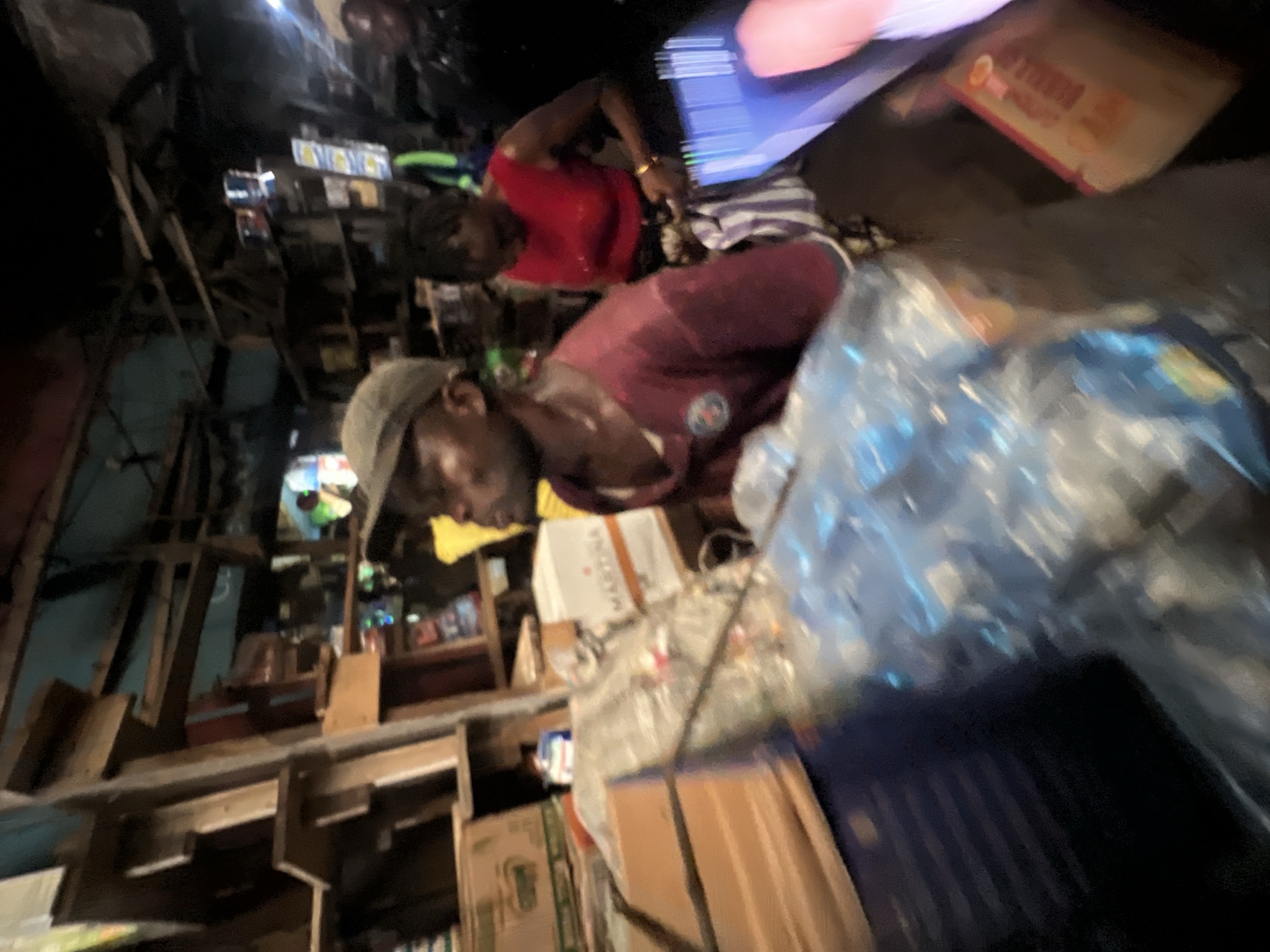 This media file captures a scene within what appears to be a market or shop in Nkpor, Nigeria. The image is dimly lit, suggesting it might be nighttime or an indoor setting with minimal lighting.
The primary subjects are two individuals. In the foreground, a person wearing a maroon shirt and a cap is leaning forward, their face partially obscured. Their expression is difficult to discern due to the low light and motion blur. In the background, another person is visible, wearing a red shirt and a gold bracelet. They appear to be interacting with something out of frame.
The setting is cluttered and appears to be an organized chaos typical of a marketplace. Shelves and wooden structures are visible, laden with various items. In the foreground, there are large plastic-wrapped bundles, possibly containing goods for sale. Several boxes and packaged items are also discernible on shelves in the background. A blue item, possibly a screen or a large piece of plastic, is prominent in the upper right portion of the frame.
The activity seems to be related to commerce or the organization of goods within a store. The overall atmosphere is that of a busy, perhaps late-night or early-morning, market. There is no clear indication of weather conditions.
Some faint text is visible on a package in the background, but it is too blurred to be legible.
