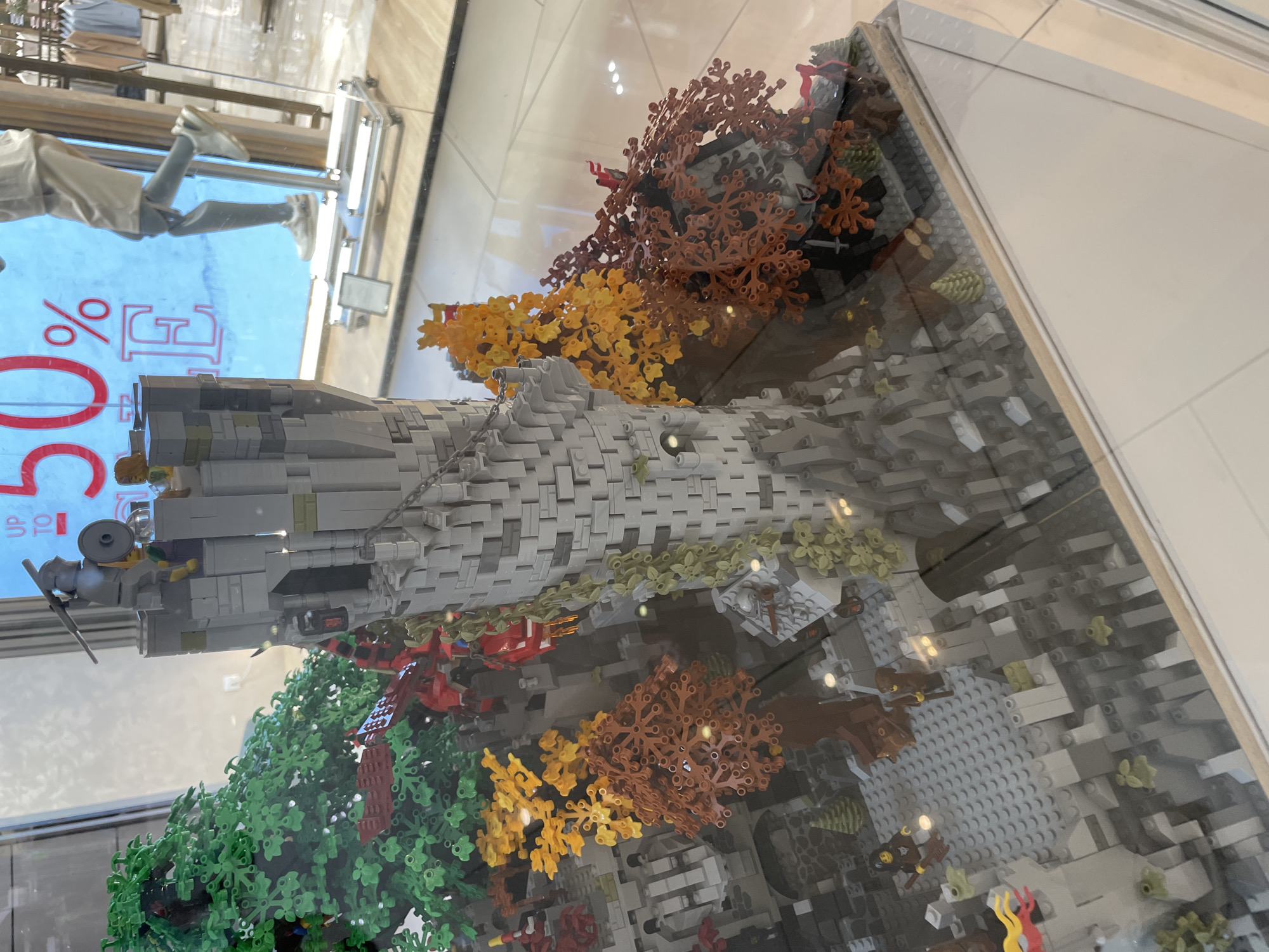 This is a close-up, eye-level shot of a LEGO model of a medieval castle and surrounding landscape, displayed behind glass. The castle is primarily constructed of gray LEGO bricks, with a tall, cylindrical tower featuring a battlemented top and a small window. A LEGO figure dressed as a knight, wearing a helmet and holding a shield and weapon, stands at the top of the tower. Several other LEGO figures, some in red armor, are depicted around the castle, suggesting a battle or siege scene.
The landscape surrounding the castle includes trees built with green, orange, and brown LEGO pieces, representing different foliage colors and seasons. There are also rocky terrain elements and a path or entranceway into the castle. The detail of the model is significant, with small LEGO elements used to create textures and features.
In the background, visible through the glass and a partially obscured display window, is a store interior. A large blue sign with red lettering reading "UP TO 50% SALE" is prominently displayed. A mannequin wearing shorts and sneakers is also visible in the store, standing on a display platform.
The overall impression is a detailed and engaging display, likely intended for a retail environment or a public exhibition. The scene appears to be a dynamic representation of a fantasy or historical setting. The specific location in Sofia, Bulgaria, is not directly discernible from the image content itself, but the presence of the store and the display suggest it is a public commercial space.