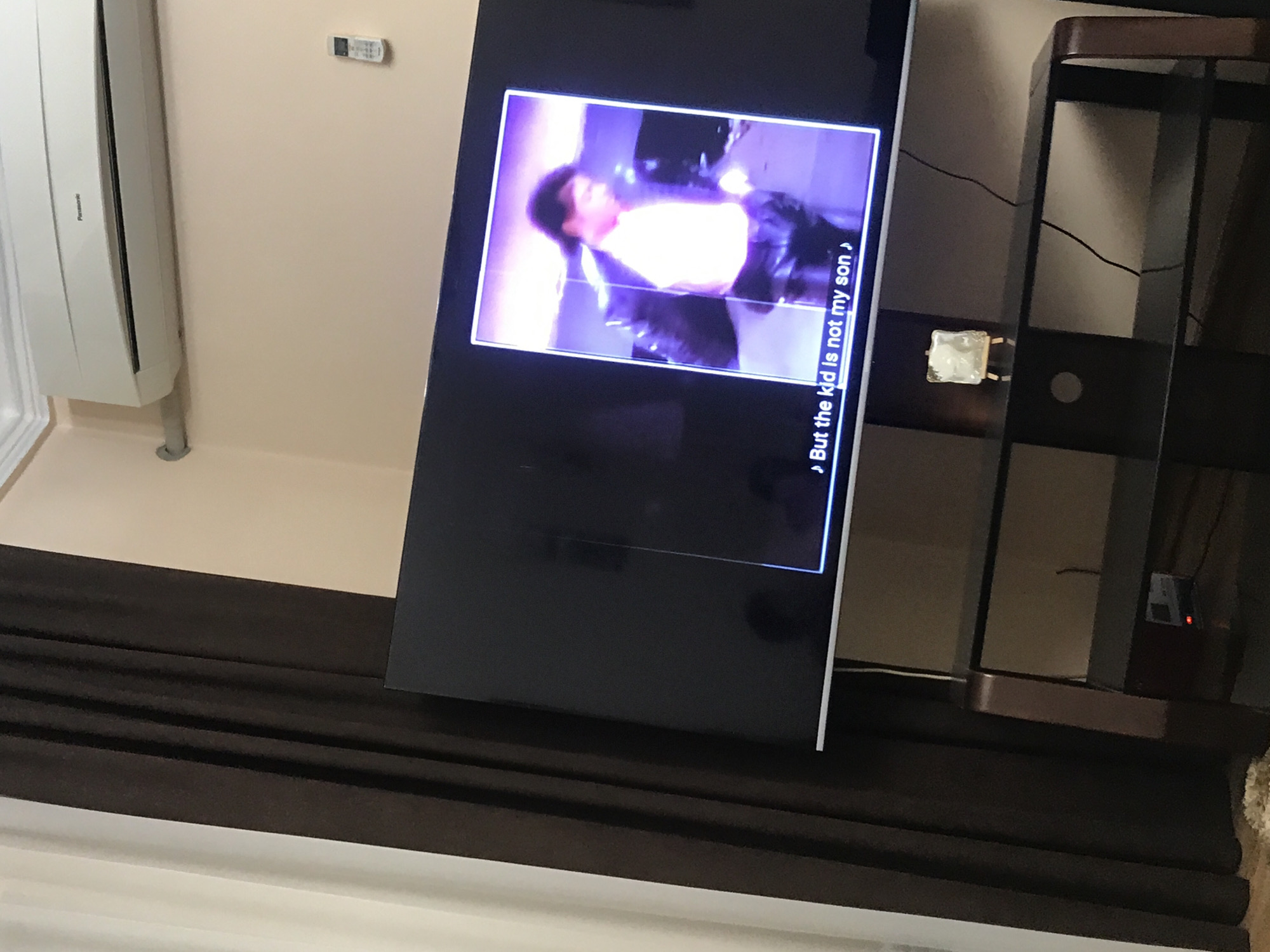 A television shows a still image of singer Michael Jackson performing, displaying the caption "But the kid is not my son". The TV is positioned in front of a dark-colored, horizontal, textured surface. Adjacent to the right of the TV is a dark-toned shelving unit. An air conditioning unit is visible in the upper left of the frame. The location cannot be determined from the image.
