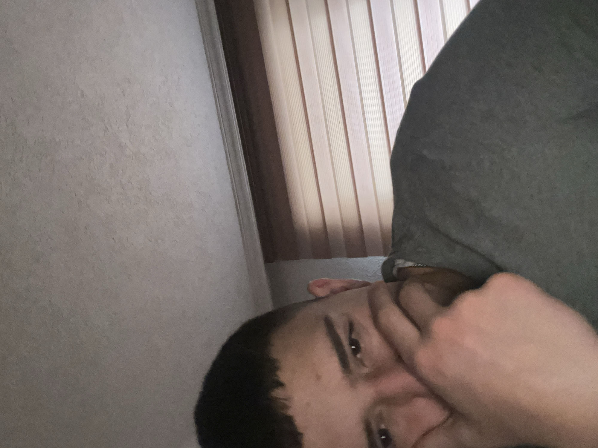 An indoor scene in what is presumed to be Zlatograd, Bulgaria, depicts a young adult male from a low-angle perspective, occupying the lower left and central portions of the frame. The male has short, dark hair and is wearing a dark grey, long-sleeved garment, possibly a sweatshirt or hoodie, with a silver chain visible around his neck. His right hand is raised, with fingers partially covering his mouth and the lower portion of his cheek. His eyes are visible, directed towards the viewer with a neutral to slight downward gaze. The upper right portion of the background consists of a light-colored window fitted with horizontal blinds, which appear to be either beige or light wood-toned. Above the window and extending across the upper left is a light-colored, textured ceiling. The lighting is internal, suggesting artificial illumination, casting soft shadows. No other subjects or objects are visible.