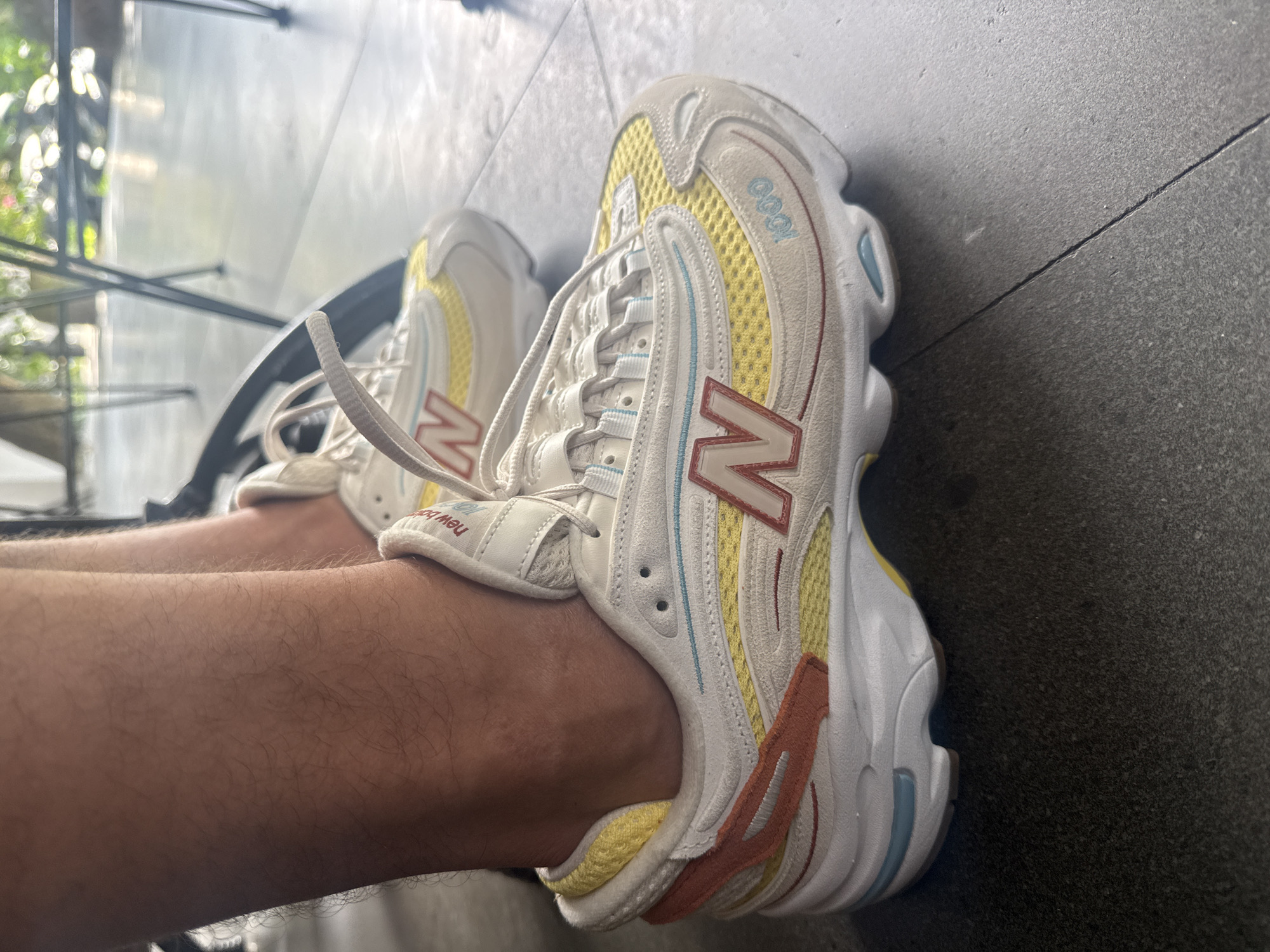 The image shows a person's legs wearing a pair of New Balance 1906R sneakers, which are predominantly white, yellow, and red with blue accents. The shoes are laced up and appear to be on a tiled, dark gray floor. The setting looks like an outdoor area, with glimpses of foliage and what might be a metal structure in the blurred background. The location is suggested to be Ubud District, Indonesia, though this cannot be definitively confirmed from the visual data alone.