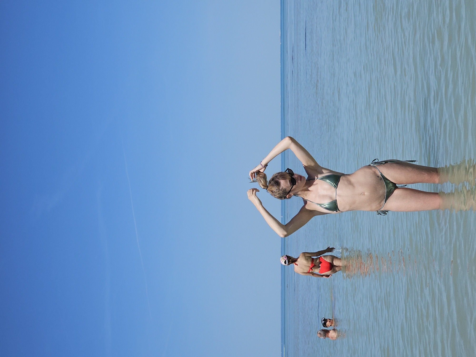 The image shows a sunny beach scene, likely in La Brée-les-Bains, France. The sky is clear blue. The water is light blue and shallow. A woman wearing a shiny green bikini is standing in the water, with her arms raised. In the background, two other people are visible in the water, one wearing a red bikini and a floral head covering, and another, further back, whose attire is not clear, but who appears to be snorkeling.