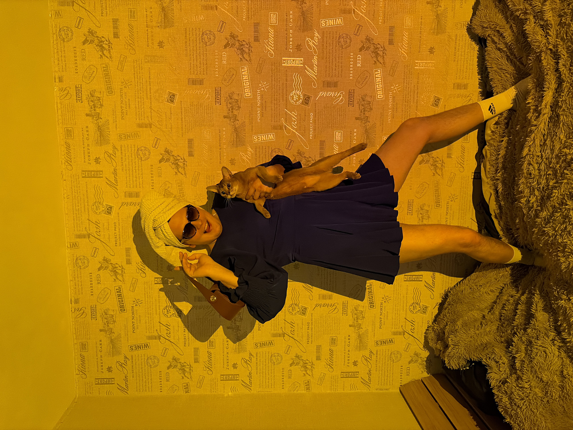A person, presumably female, is lying on their back on a surface with a tan and brown patterned wallpaper. The surface appears to be a bed. The person has a towel wrapped around their head, sunglasses on their face, and is wearing a dark blue dress. The person is holding a brown cat on their body. The cat is reddish-brown and appears to be relaxed. The person is wearing white socks with black Adidas stripes. The room appears to be lit by a warm, yellowish light. The patterned wallpaper has text and graphic design elements, including the words "Kiona" "Martin Ray" and "Joseph Carr" along with "Wines". The location information provided, Chaikovsky, Russia, is external and not discernible from the image itself.