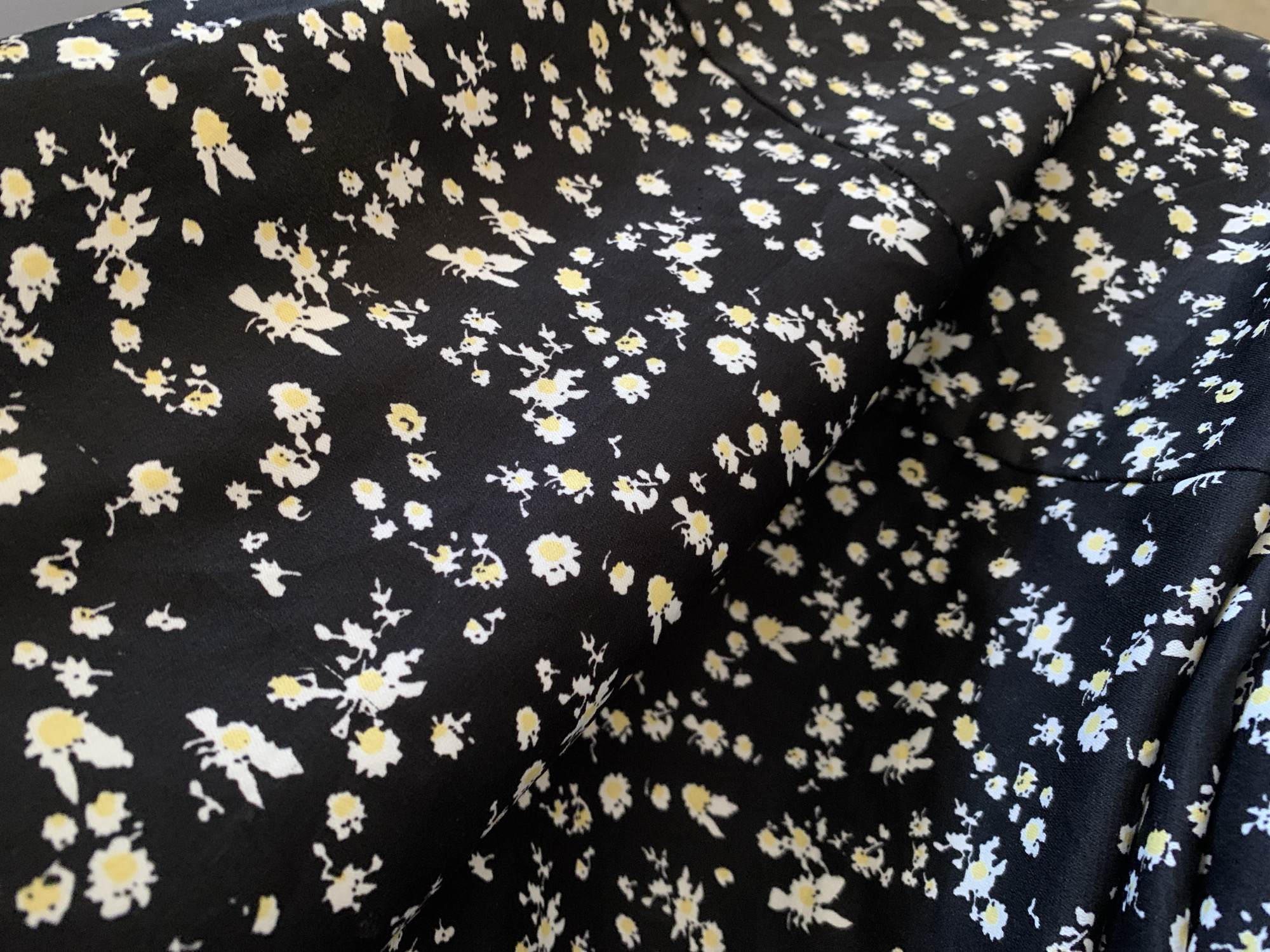 A close-up shot captures a black fabric with a delicate, scattered floral print. The material is folded and draped, creating soft contours and shadows that emphasize its flow and texture. The background of the fabric is a solid, deep black. The floral pattern features numerous small, stylized blossoms, each with white petals and soft, pale yellow centers, resembling tiny daisies or wildflowers. These small flowers are randomly distributed across the textile, creating a gentle and intricate design.
The fabric appears soft and pliable, suggesting a light material like chiffon or rayon. The even and diffused lighting illuminates the fabric without harsh reflections, allowing the details of the print and the subtle texture to be clearly visible. No people or other objects are present in the frame. There is no discernible activity taking place, as the image focuses solely on the static fabric. The setting is not explicitly clear beyond the textile itself, with no visible cues for time of day, weather, or specific location. There is no visible text in the image. The visual content does not offer any information related to the external location context of "New Stadium, Nigeria."