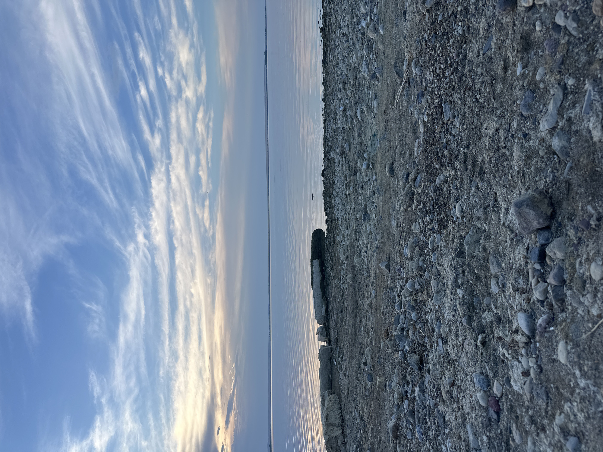 The image depicts a serene natural landscape at dusk, likely near the city of Energetik, Russia, given the presence of a vast body of calm water, characteristic of the landscape surrounding the city, which is known for its proximity to Lake Baikal and its hydroelectric power station.
The foreground is dominated by a rocky, pebble-strewn shore. The pebbles are varied in size, shape, and color, ranging from small grains of sand to larger, rounded stones in shades of gray, brown, and white. Some larger, weathered concrete-like blocks are scattered along the shoreline, appearing to be man-made debris or remnants of structures. A thin layer of sand is mixed with the pebbles in some areas.
Beyond the shore lies a vast expanse of tranquil water, reflecting the soft, diffused light of the twilight sky. The water's surface is smooth, with subtle ripples creating a shimmering effect. A clear horizon line separates the water from the sky.
The sky is a prominent feature, painted with soft hues of blue, gray, and faint yellow. Wispy, elongated clouds stretch across the upper portion of the frame, creating a delicate, feathery pattern. The light suggests it is either early morning or late evening, with the sun dipping below the horizon or just beginning to rise. No people are visible in the image, and the overall impression is one of quiet solitude and natural beauty.