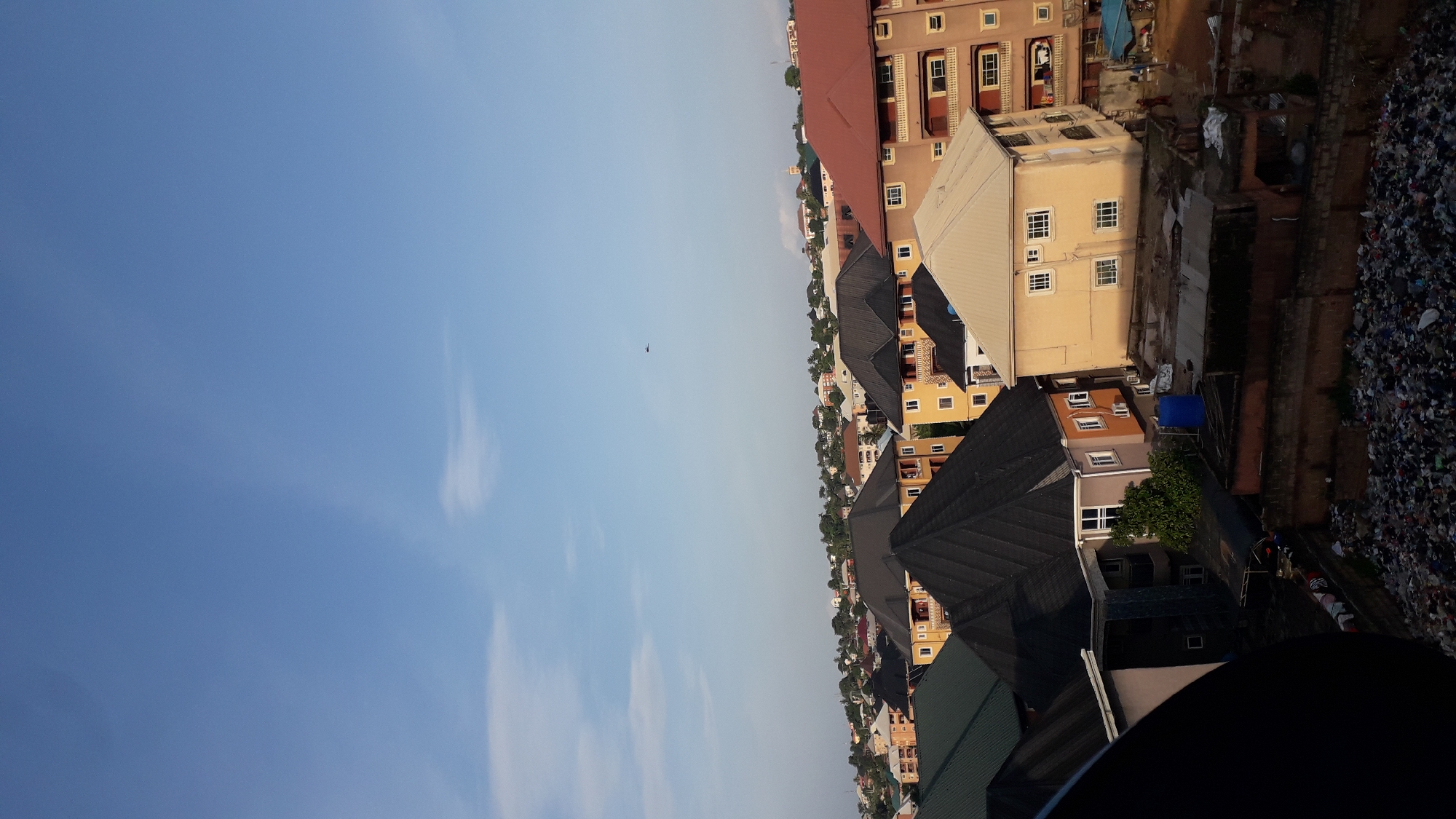 The image captures a view of a dense urban area, likely a residential neighborhood, with numerous buildings and a clear blue sky. The architecture suggests a developing city, with a mix of multi-story residential buildings and some older structures. Several houses feature distinct roof designs, with prominent gables and varied color palettes. In the foreground, a large pile of accumulated refuse is visible, indicating a sanitation issue in the area. A dark, curved object, possibly a satellite dish or part of a camera, is partially visible in the lower left corner, suggesting the vantage point from which the photo was taken. In the distance, more buildings and greenery are discernible, stretching towards the horizon. A tiny object, potentially an aircraft, is visible in the upper portion of the sky. The overall scene depicts a bustling, possibly lower-income urban environment.