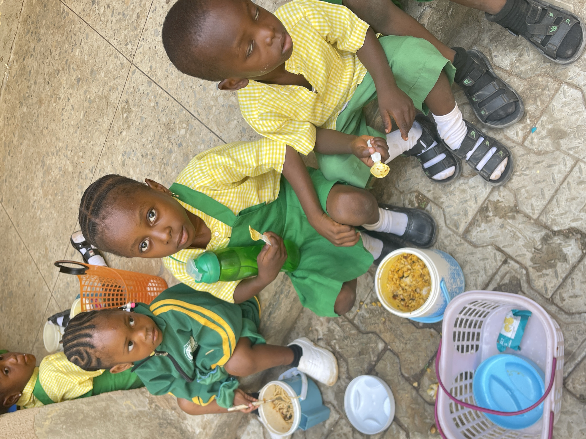 The image depicts three young children in school uniforms seated on a patterned, light-colored surface. The uniforms consist of green and yellow combinations, with one child wearing a yellow checkered shirt and green shorts, another wearing a similar shirt with a green dress, and the third wearing a green jacket with yellow stripes and matching green shorts. The children appear to be eating lunch from containers and using utensils. Several lunchboxes, containers, and a basket are scattered around them. The location is presumed to be outdoors, possibly within a schoolyard or similar area, and identified as Akwanga, Nigeria. The scene suggests a lunchtime setting for school children.
