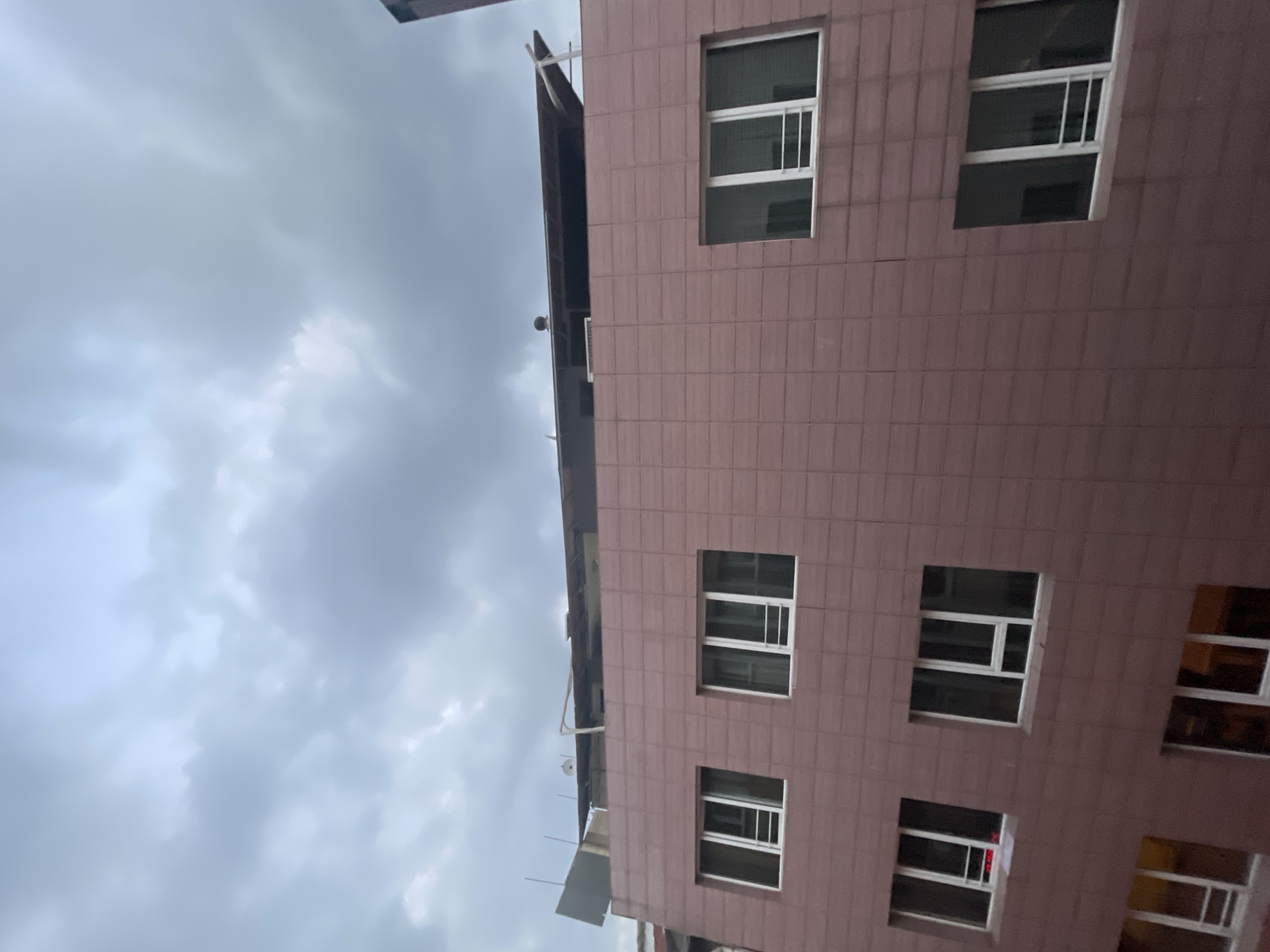 The image shows the exterior of a multi-story building with a reddish-brown tiled facade under a cloudy sky. The building has several windows with white frames. On the top of the building, there is a rooftop structure with some additional rooms or extensions and exposed roofing materials. The sky is overcast with gray clouds. The lower portion of the image shows more windows and the continuation of the tiled facade. The scene appears to be an urban environment, possibly in Kadıköy, Turkey, given the architectural style and common building materials. There are no people or visible actions taking place in the image. The perspective is from a slightly elevated viewpoint, looking upwards at the building.