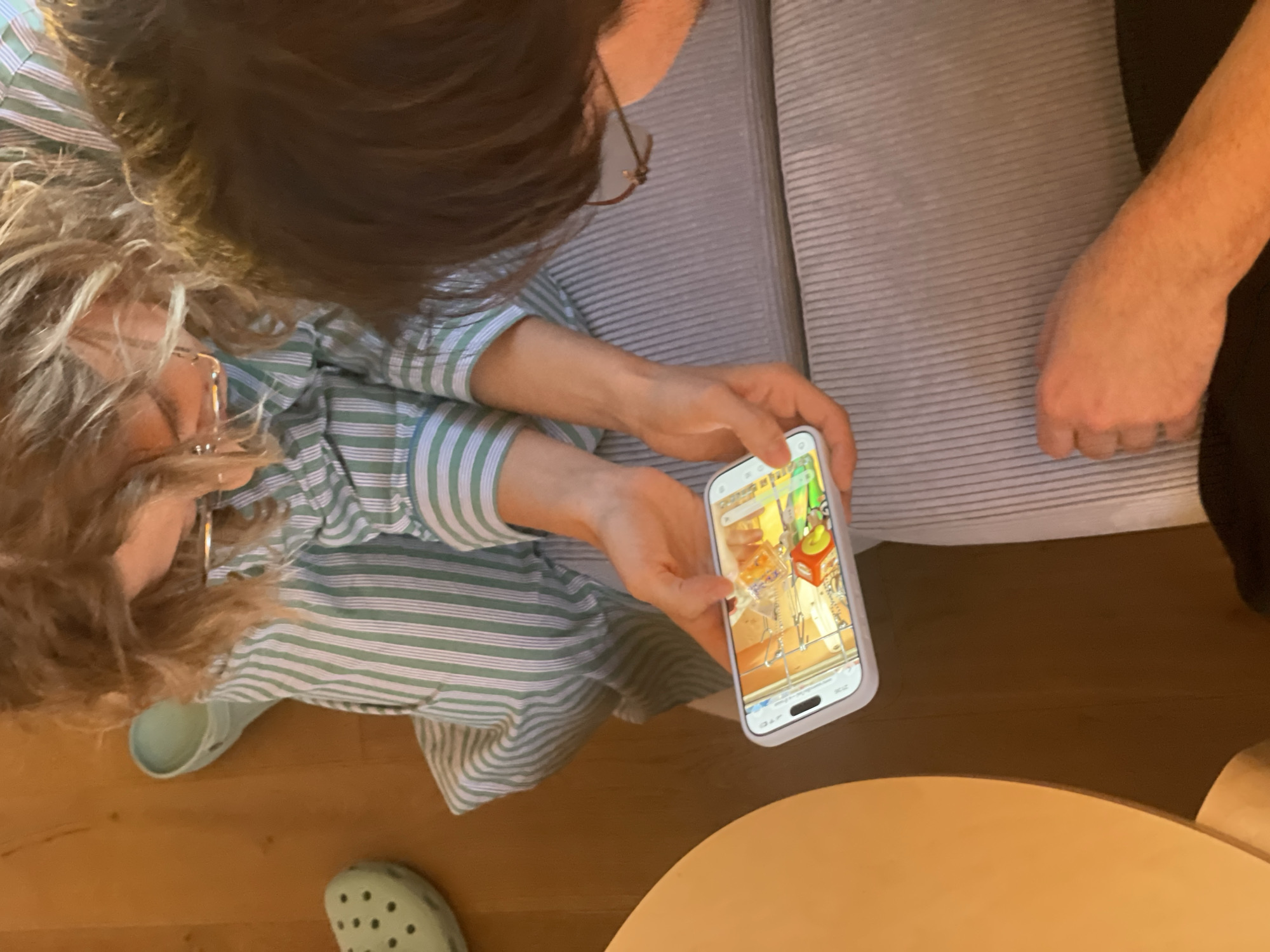A high-angle shot captures two people sitting indoors. One person, wearing a light green and white striped shirt and glasses, is holding a smartphone displaying an image, while wearing light green crocs. The other person, wearing glasses, sits next to them. Both are seated on what appears to be a grey corduroy couch. The environment includes a wooden floor, and a round, light brown table. The image does not contain enough details to confirm the location as Saint Petersburg, Russia.
