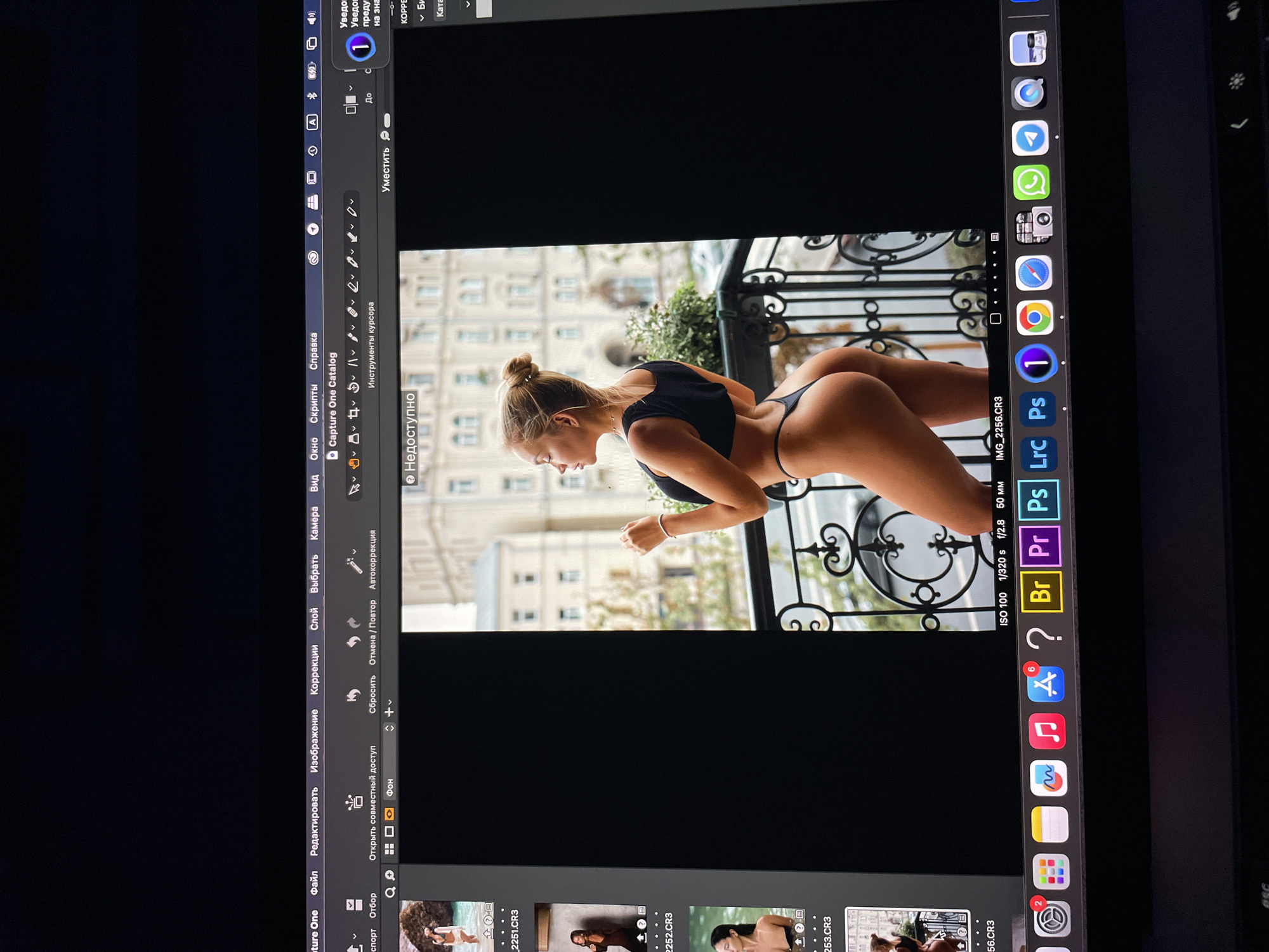 A computer screen displays an image of a woman in a black top and thong, bending forward on a balcony with an ornate railing. Behind her is a building facade with multiple windows. The photo is open in Capture One editing software. The computer's dock on the right shows icons for various apps like Safari, Chrome, Photoshop, and Final Cut Pro. The software interface is in Russian. The location is likely in Kazan, Russia, due to the Cyrillic text and architectural style.