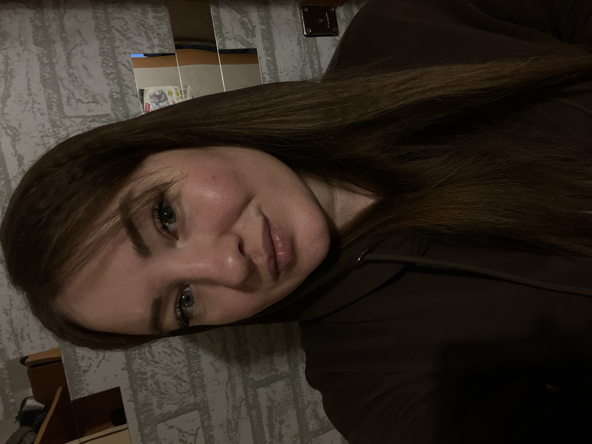A woman with long brown hair and blue eyes is posing for a selfie in a room. The background appears to be a wall with a textured pattern resembling brick or stone. There's a reflection of a mirror and furniture in the background, suggesting she's indoors. The woman is wearing a dark brown hooded sweatshirt. The image does not contain enough information to determine the location in the city of Chaikovsky, Russia.