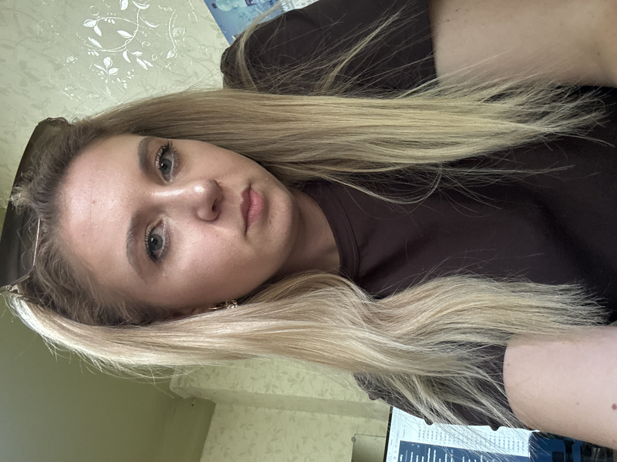 This is a close-up, low-angle, portrait-style photo of a young woman with long, blonde hair, taken indoors. The woman is looking directly at the camera, with her head tilted slightly. Her eyes are open and she appears to be making eye contact with the viewer. Her hair is a dominant feature, falling around her face and shoulders, and appears to be slightly tousled. She is wearing a dark-colored shirt.
The background is slightly out of focus, but appears to be a domestic or office setting. There is a wall with a subtle floral pattern behind her, and to the right, a laptop screen displaying text and what looks like a spreadsheet or table. The lighting suggests it is daytime, but there are no specific cues for time of day or weather. The woman's expression is neutral, and there are no strong emotional indicators.
Visible text on the laptop screen is partially obscured and difficult to read clearly, but includes dates and what appear to be entries in a list format. Some of the readable text includes dates like "1.06.26" and "17.06.26".