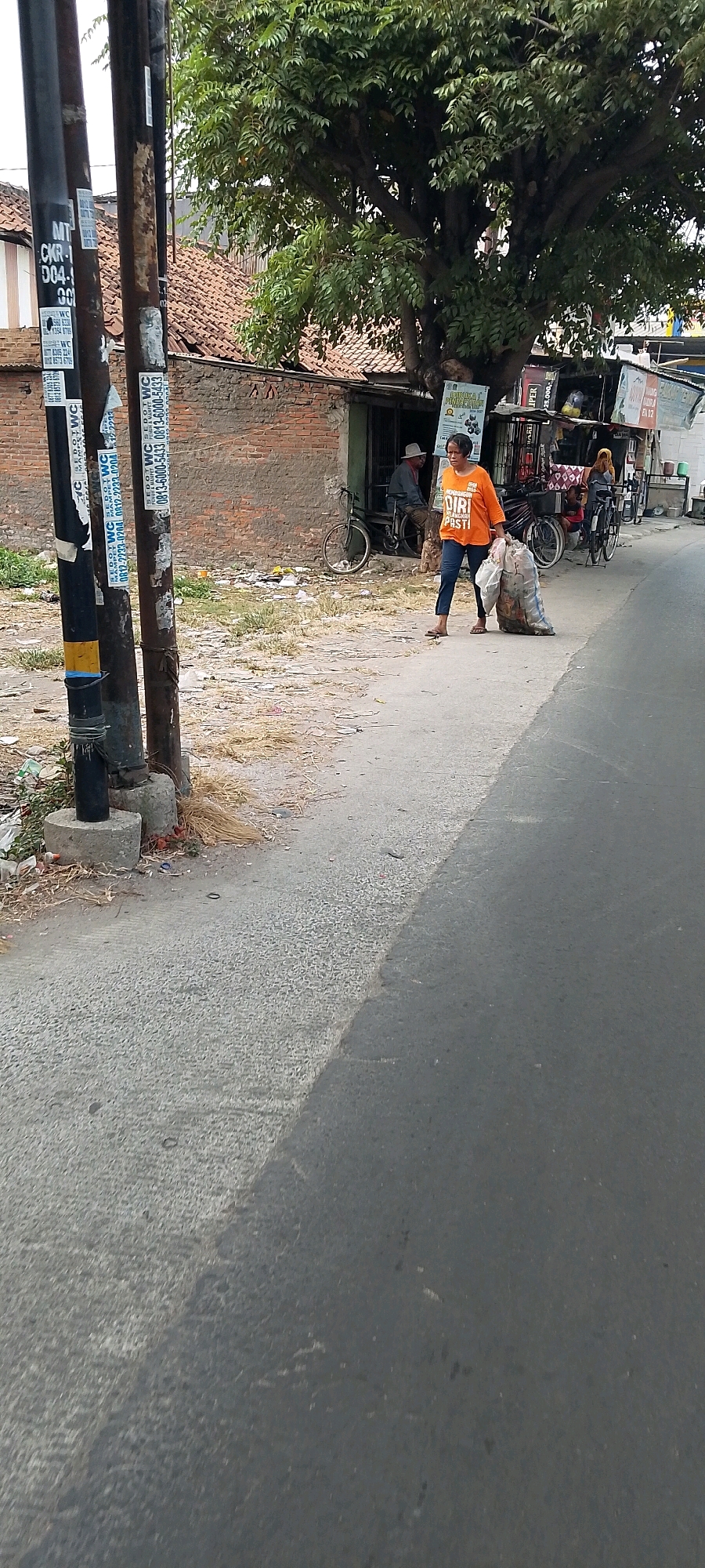 An outdoor medium shot captures a woman in an orange t-shirt and dark pants walking along a street in Satriamekar, Indonesia, carrying a large, bundled bag of trash. She is walking from left to right across the frame. The street is paved with asphalt, with a narrower section of lighter-colored textured concrete on the left. Scattered debris, including plastic bags and dry vegetation, lines the edge of the concrete. A dark metal pole with various stickers, possibly advertisements or service notifications, stands on the left side of the image, with concrete bases. In the background, a brick building with a corrugated metal roof is visible, partially obscured by a large tree with dense green foliage. To the right of the tree, a modest storefront or stall displays signs and various items. Two bicycles are parked near the storefront. Another person, partially visible, sits on a bicycle in the background, facing away from the camera. The scene suggests a residential or commercial area in a moderately populated Indonesian town.