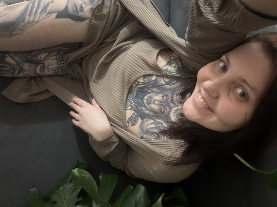 A woman smiles at the viewer, reclining on a black surface. She wears a ribbed, khaki-colored sweater or dress. Visible tattoos adorn her chest, arms, and legs, featuring faces and serpent-like designs. Part of a green houseplant, possibly a Monstera, is visible in the foreground. The image is taken from a slightly high angle, capturing the woman's relaxed posture and the details of her tattoos.