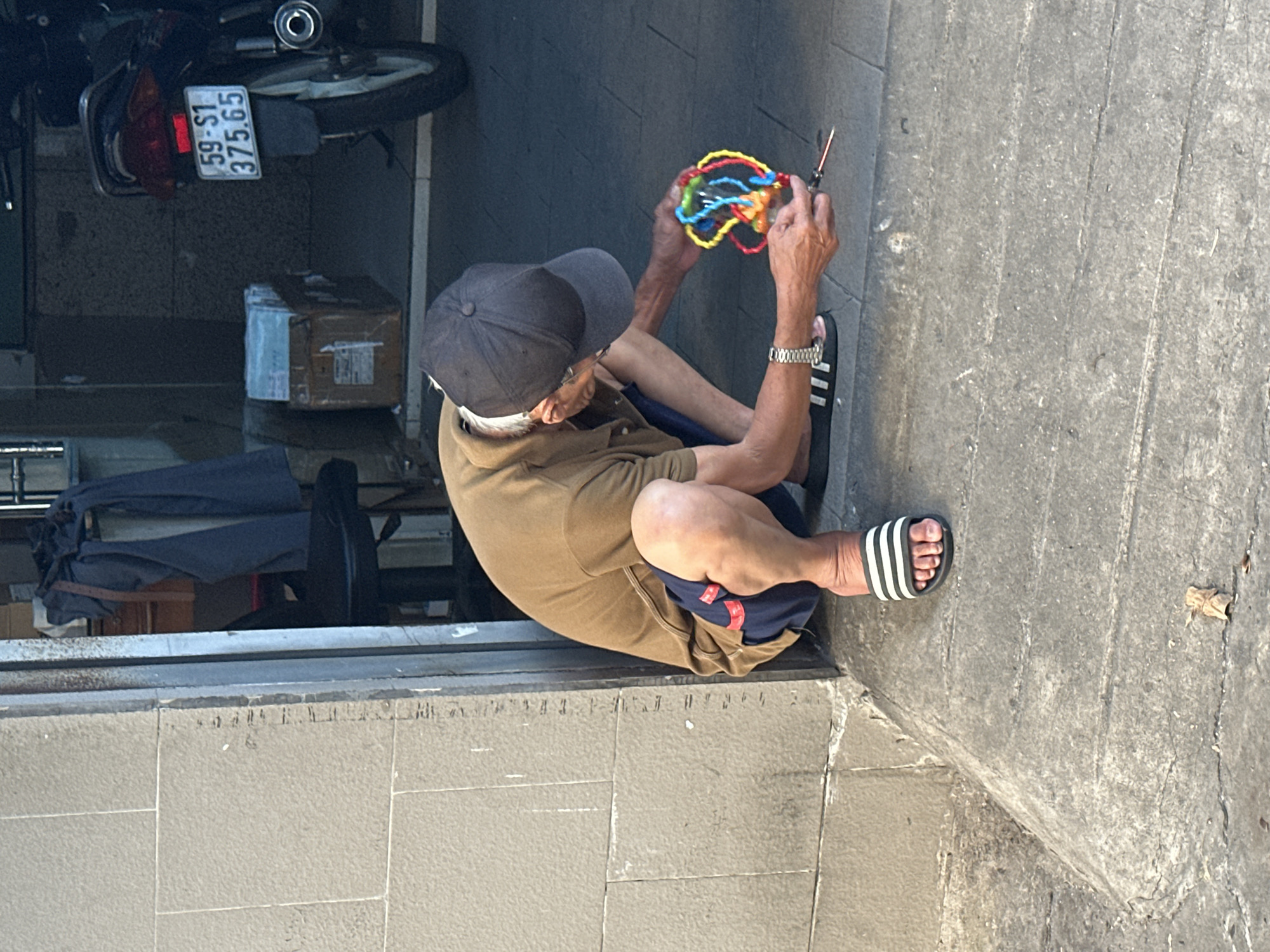 A man wearing a brown shirt, dark shorts, a dark baseball cap, and Adidas-striped sandals sits cross-legged on a grey concrete sidewalk in Thủ Đức, Vietnam. He is working with a colorful, abstract object made of wire or thread, manipulating it with a small tool. A motorcycle is parked behind him, showing a license plate with "59-S1 375.65". There are boxes and clothes visible behind the man. The background shows a typical urban street scene.