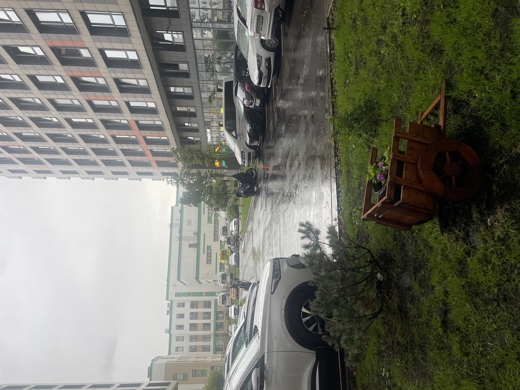 In the city of Poroshkino, Russia, on a cloudy day with light rain, a parking lot is wet and reflects the overcast sky. Several cars are parked in neat rows, including a white SUV in the foreground, a black sedan, a white sedan, and a black motorcycle covered with a tarp. To the right of the parking lot, a modern apartment building with numerous windows and an orange accent design stands tall. Across the street, a row of buildings, including one with a green trim, is visible, and a yellow ambulance is parked among other vehicles. In the foreground, a small evergreen shrub sits on a patch of grass, and to its right, a rustic wooden cart filled with flowers serves as a decorative element. The grass is wet, with some muddy patches visible, indicating recent rainfall.