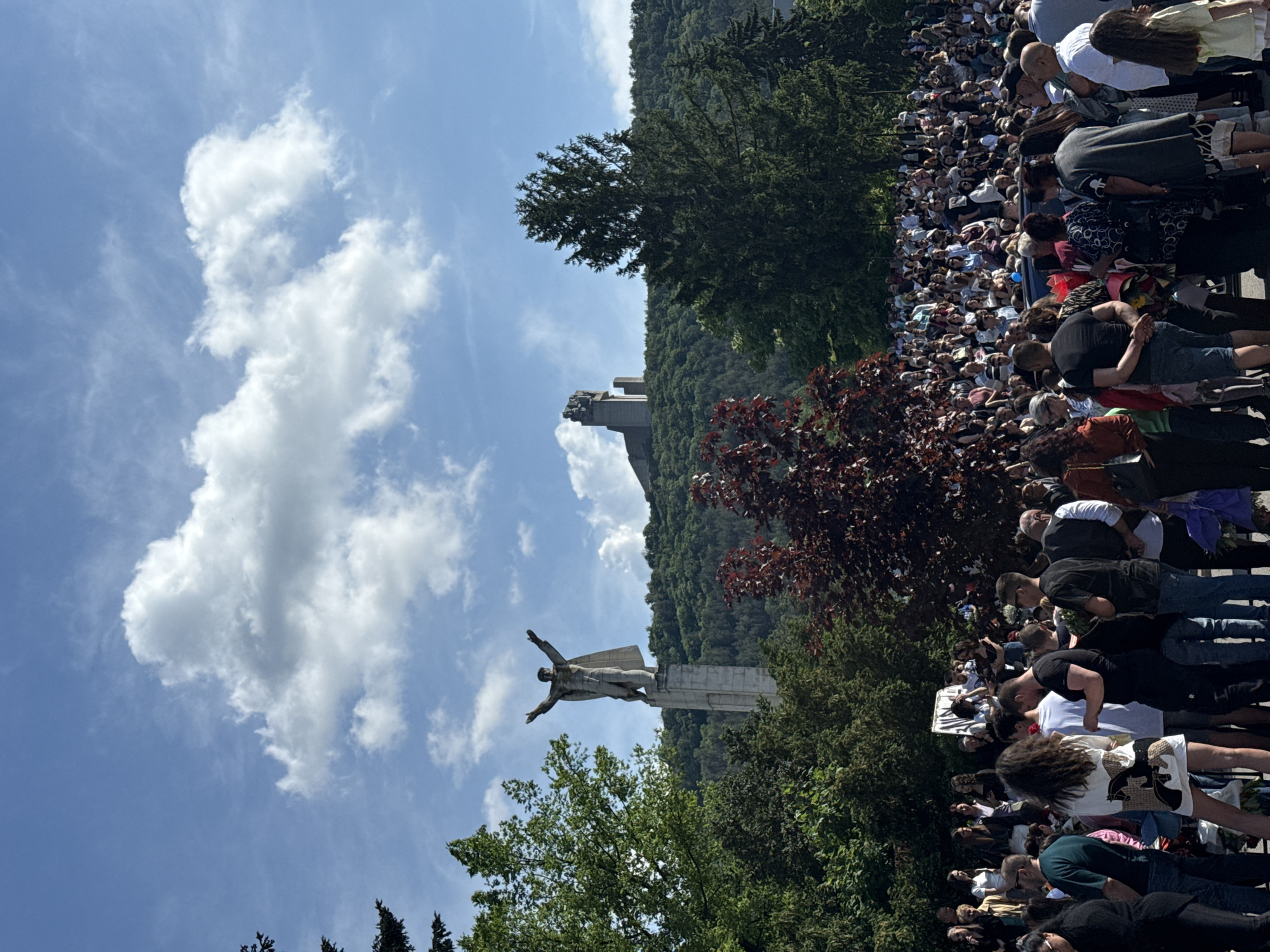 A crowd of people is gathered on a sunny day near the Monument to 1300 Years of Bulgaria in Shumen, Bulgaria. The monument, featuring a large concrete statue of a man with outstretched arms, stands on a hill in the background. The sky is blue with some white clouds. There are trees and vegetation in the foreground and on the hill.