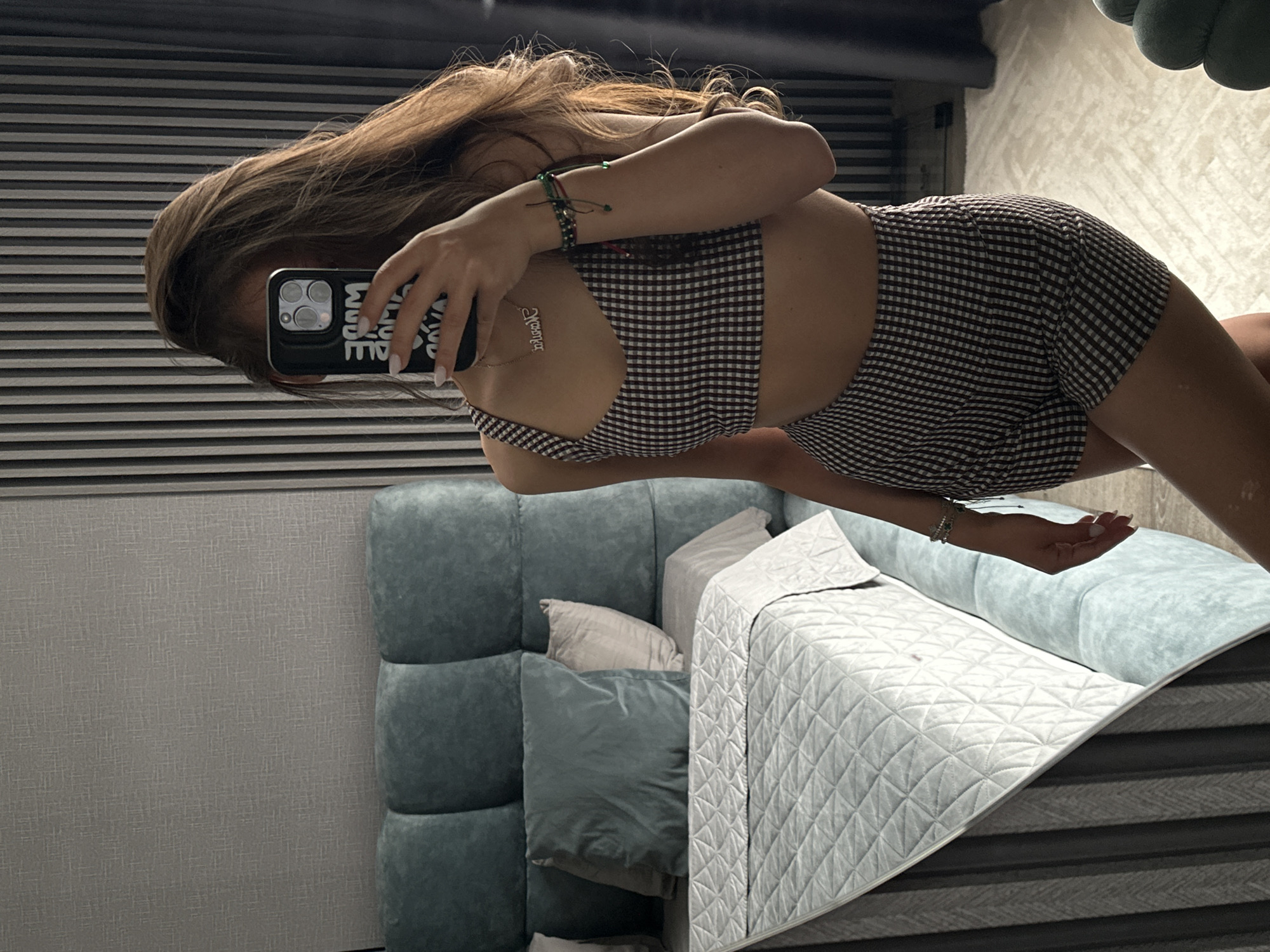 A young woman is taking a mirror selfie in a bedroom. She is wearing a checkered crop top and shorts. The bedroom has a large bed with a teal headboard and light-colored bedding. The walls are decorated with textured wallpaper and vertical striped paneling. The woman is holding a phone in her right hand, with a phone case that has bold lettering. She is also wearing a necklace and bracelets. The overall impression is of a modern, stylish bedroom setting.