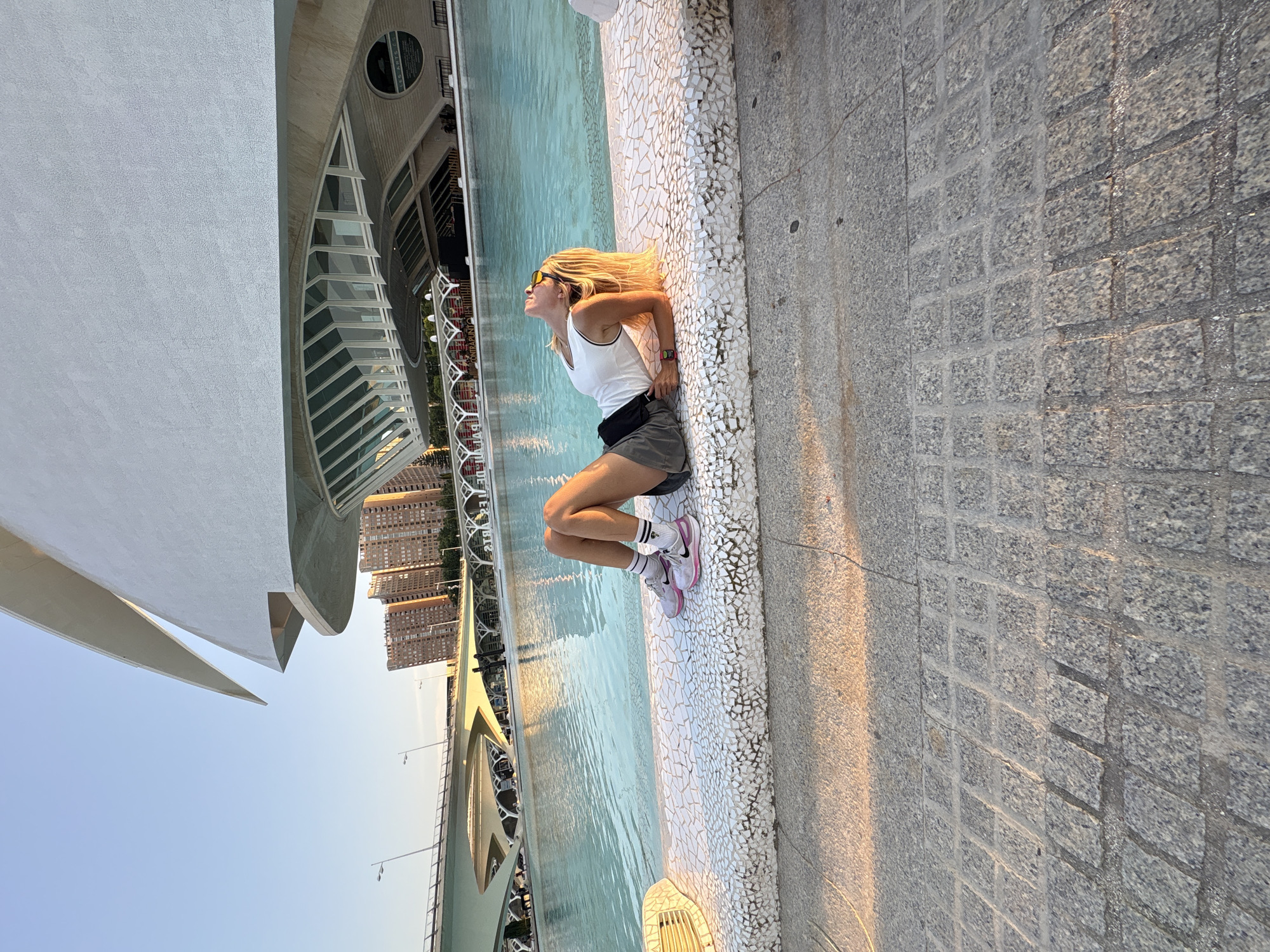 A woman with blonde hair and sunglasses is lying on a textured concrete ledge next to a large body of turquoise water. She is wearing a white tank top, grey shorts, and pink sneakers with white socks. Her head is tilted back, looking up at the sky. In the background, a modern white building with a distinctive arched roof and large windows overlooks the water. A bridge with arched supports and tall buildings are visible further in the distance. The scene appears to be in a public space in the City of Arts and Sciences in Valencia, Spain, possibly the Hemisfèric building, given the architectural style. The ground in the foreground is paved with large, gray granite stones, and the lighting suggests it is late afternoon or early evening, casting long shadows.
