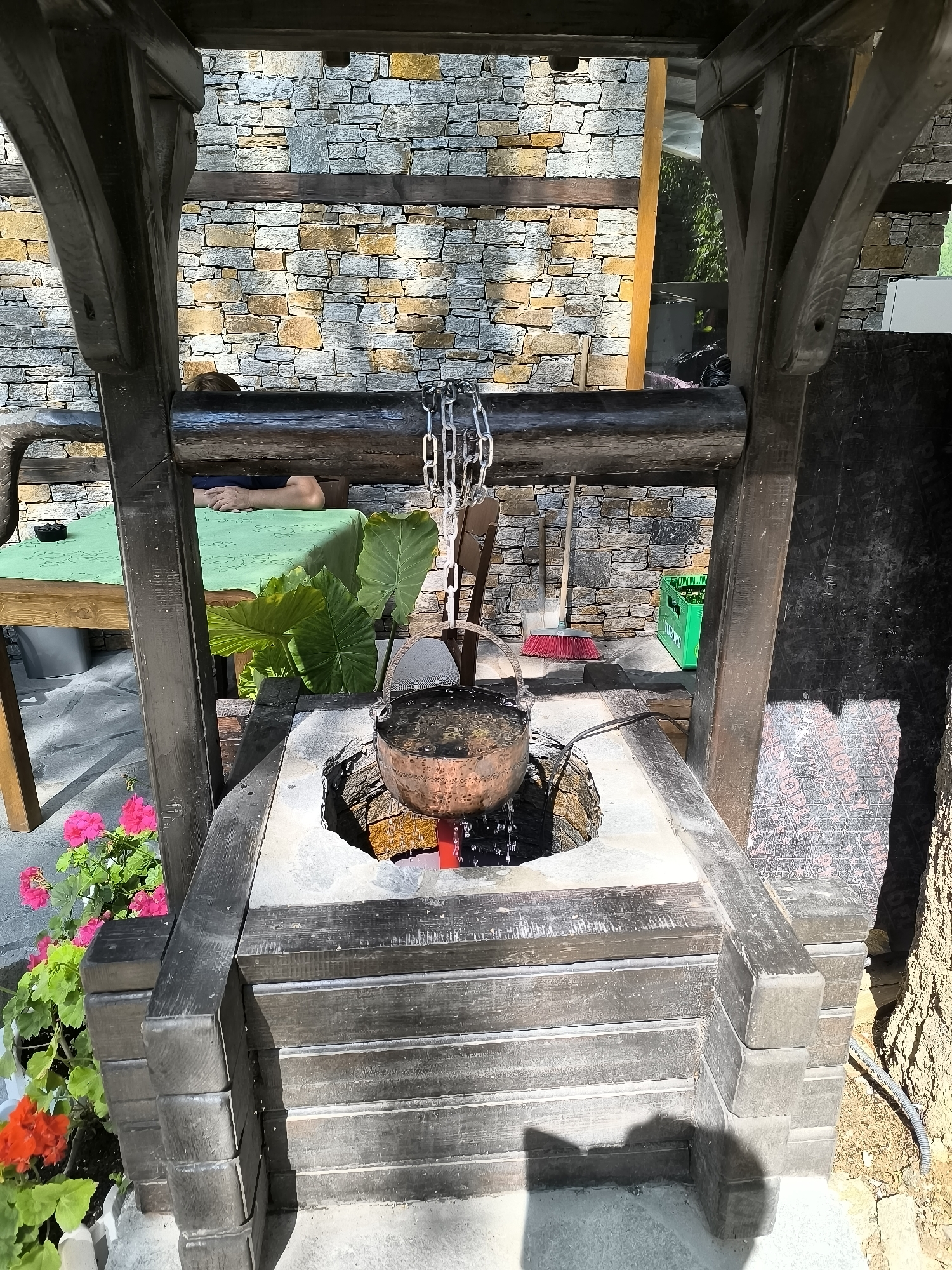 The image shows an outdoor scene in Kovachevitsa, Bulgaria, centered around a well structure. A copper pot filled with water is suspended over an opening in the well, with a chain attached to a wooden beam above. Water is visibly dripping from the pot. The well is constructed with dark wooden beams and stone blocks. In the background, a stone wall provides a textured backdrop. To the left, a person is partially visible, resting on a green tablecloth-covered surface. A table, large green leaves, a broom with a red brush, and a green crate filled with bottles are also present. Pink and red flowers are in the foreground, planted in pots. The lighting suggests it is daytime, with shadows cast on the ground.