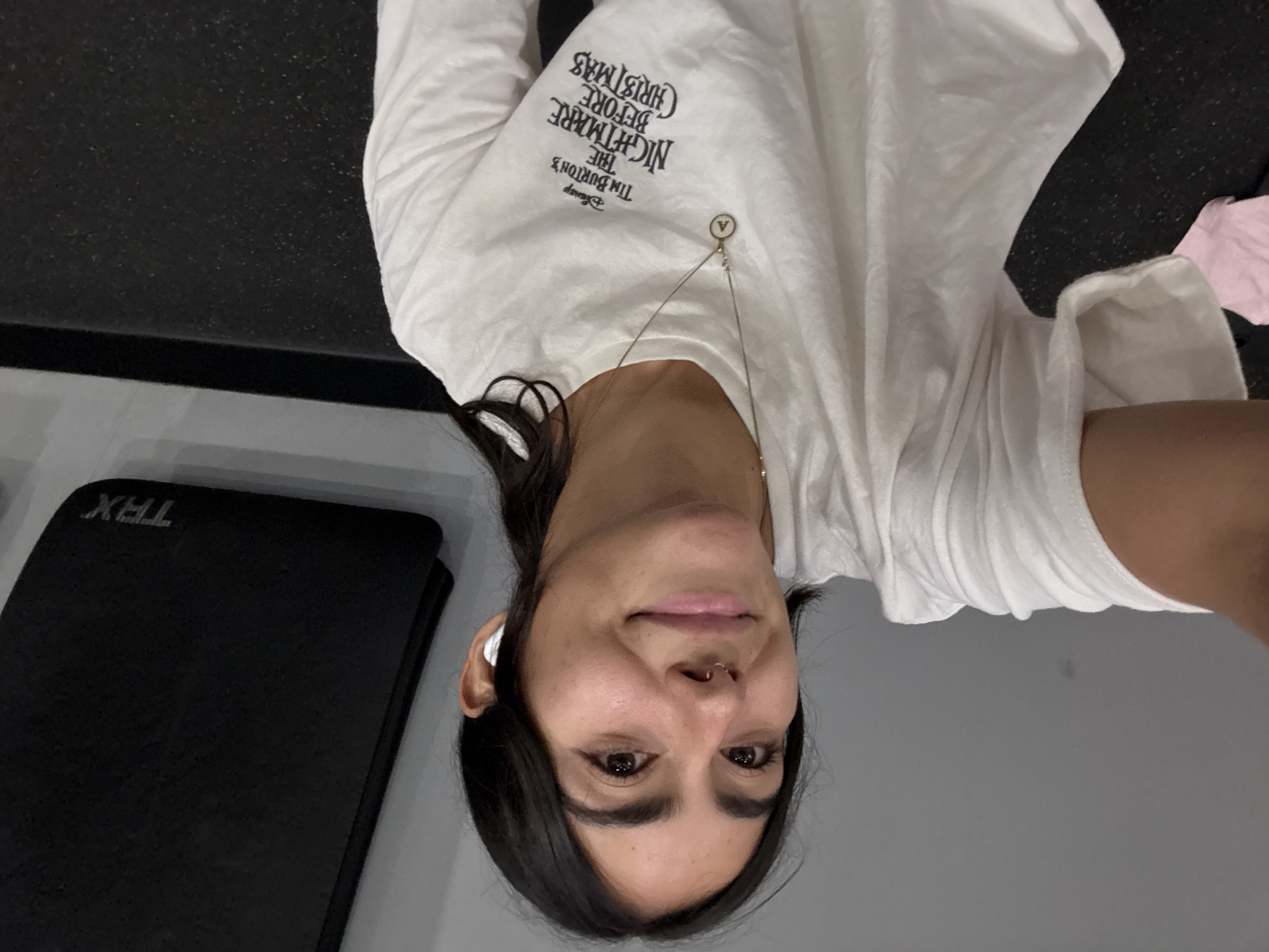 A medium-angle, indoor selfie depicts a woman with dark hair, wearing a white long-sleeved t-shirt featuring "The Nightmare Before Christmas" print and a gold necklace, with a visible nose ring and a white earbud in her left ear. A black TRX mat is visible to the left of the frame, and a grey wall is in the background. The woman appears to be in a gym-like setting, although the specific location within the city of Puebla, Mexico is not discernible from the image. There are no visible interactions or actions occurring.